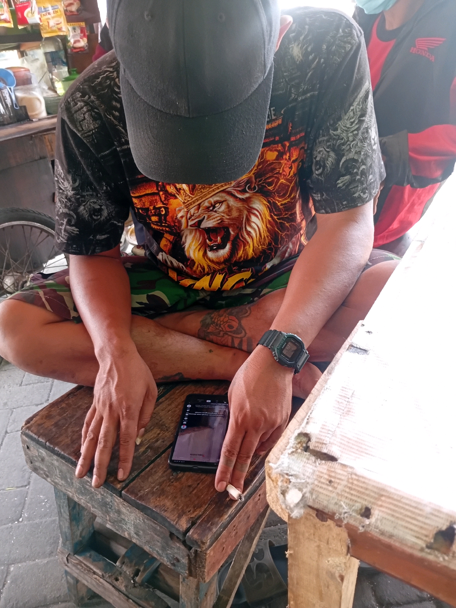 The image depicts a close-up view of a person sitting on a rustic wooden stool, with their legs crossed. The person is wearing a black baseball cap and a t-shirt featuring a prominent graphic of a lion's head. They are also wearing camouflage shorts. In front of them, on a weathered wooden table, lies a smartphone, and the person's right hand is holding a lit cigarette. The background suggests an outdoor setting, possibly a street-side stall or warung, with glimpses of shelves stocked with various items and a bicycle wheel visible to the left. The overall scene has a casual and unpretentious feel. It is highly probable this is in the city of Malang, Indonesia, given the presence of the warung style and local ambiance.