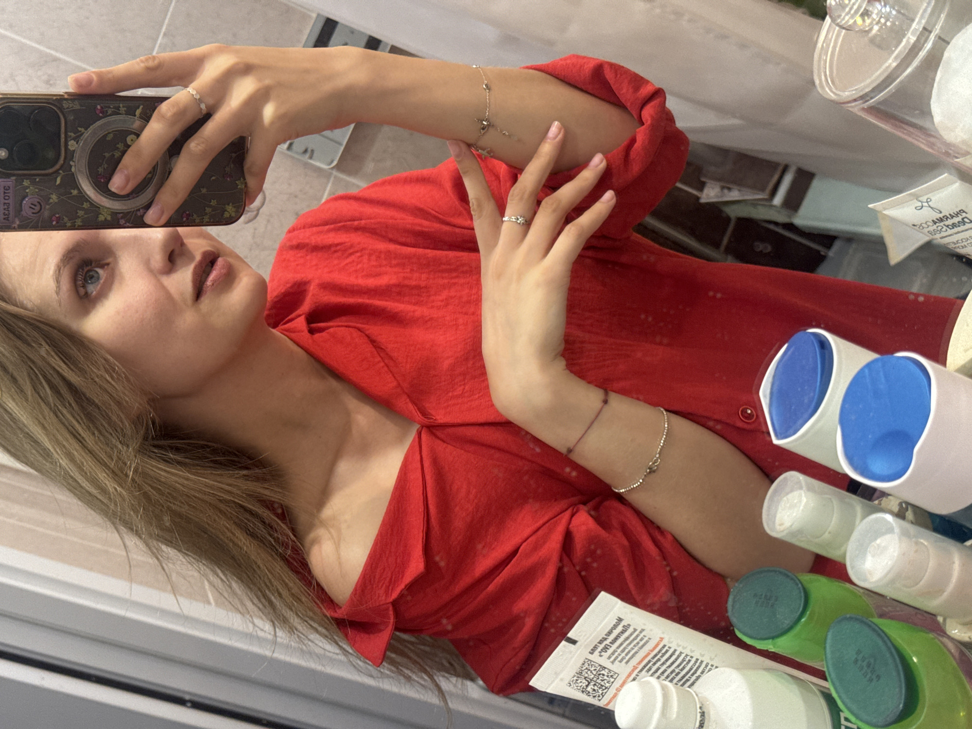 A woman is taking a selfie in a bathroom mirror. She is wearing a red shirt. Her phone has a floral design on the case. She is wearing rings and bracelets. In the background, there are toiletries such as bottles of lotion and shampoo, along with cosmetics and hair products. The specific location in Izhevsk, Russia, is indeterminable.