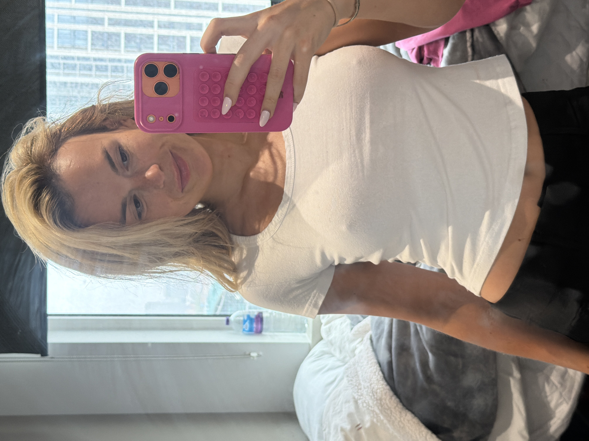 A blonde woman takes a mirror selfie in a room. She is wearing a white t-shirt and black leggings. She is holding a pink phone with a camera, and has manicured nails. Behind her is a bed with grey and white blankets. A window is visible behind the bed, with a water bottle on the windowsill. The scene appears to be an apartment in London, United Kingdom.