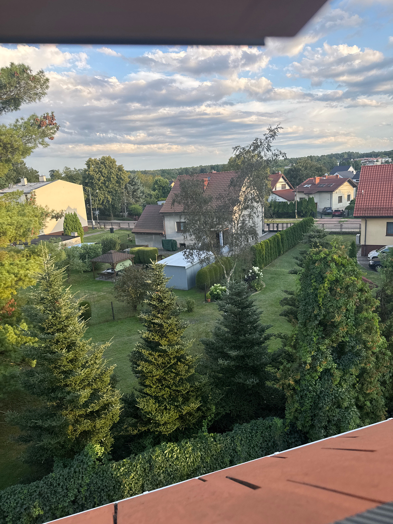 This is a vertical, medium-angle, outdoor photograph taken from a slightly elevated position, possibly a balcony, looking down onto a suburban residential area under a cloudy sky. The foreground features a portion of a reddish-brown tiled roof with some thin, dark shadows cast upon it.
The middle ground displays a well-maintained backyard with lush green grass. Several evergreen trees, including prominent fir trees, are scattered across the lawn. A wooden gazebo with a brown roof is situated in the center of the yard, appearing to be an outdoor seating area. To the left of the gazebo is a light blue metal shed. A neat row of tall, dark green hedges runs diagonally across the yard, separating it from neighboring properties. Beyond the hedge, more houses with red tile roofs and white or light yellow walls are visible. One house in the background has a large, light yellow facade. A few parked cars are visible on the street behind the houses.
The background consists of a dense line of trees forming a natural barrier, with more distant houses and buildings beyond them, suggesting a relatively spread-out suburban landscape. The sky is partly cloudy with patches of blue sky visible, indicating it's likely daytime, possibly late afternoon given the angle of the sun casting shadows. The overall impression is one of a quiet, residential neighborhood on a pleasant day. The image does not contain any people or any explicit indication of the city of Katowice, Poland, beyond what has been specified in the prompt.