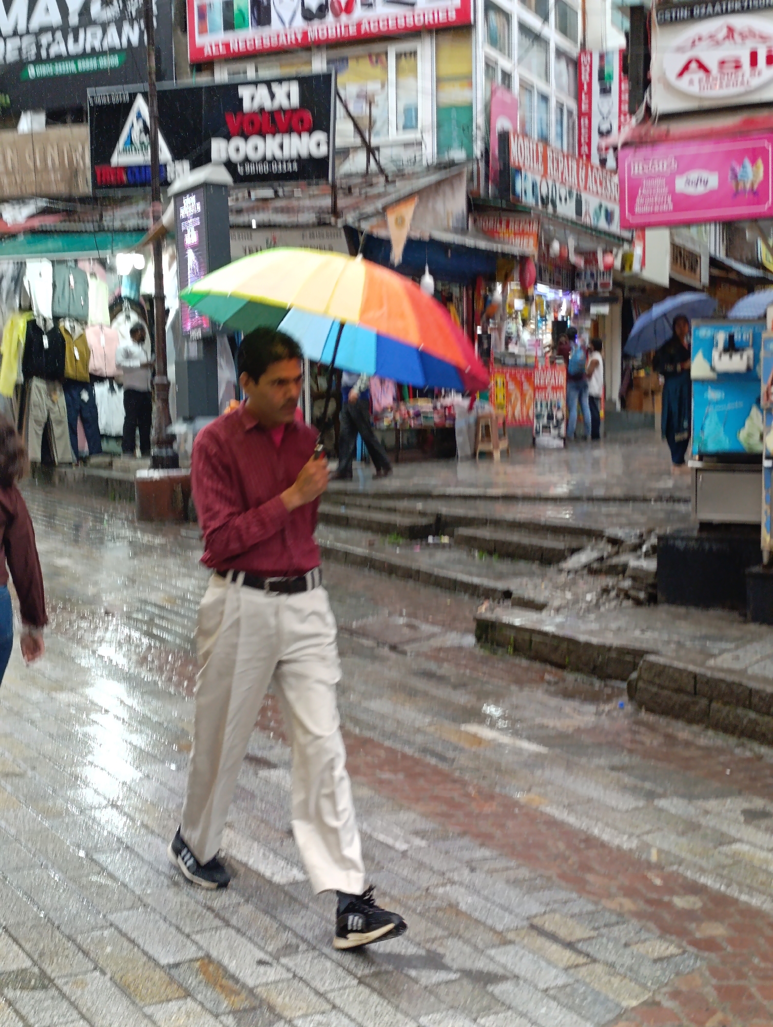 In Manali, India, a man with dark hair, wearing a red collared shirt and light-colored pants, walks with a rainbow-colored umbrella open above him. The ground is wet and reflective, indicating recent rain. The man is wearing black athletic shoes with white accents. He is walking on a paved street lined with shops. Behind him, a colorful awning is visible, and further back, shops display various goods. Signs for "TAXI VOLVO BOOKING" and "MOBILE REPAIR & ACCESSORIES" are visible. The overall scene depicts a busy street during or immediately after rainfall.