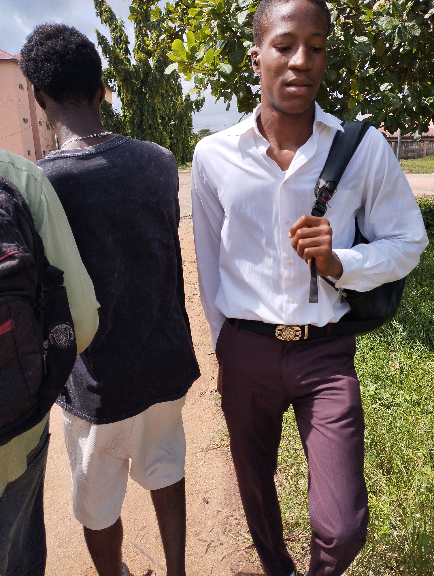 Three young African men are standing outdoors on a dirt path in Ihiagwa, Nigeria. The man on the right wears a white button-down shirt, dark maroon trousers, a black belt with a gold buckle, and has a black backpack slung over his left shoulder. He is also wearing a black earbud in his left ear. His gaze is directed downwards.
To his left, another man is seen from the back, wearing a dark gray t-shirt and white shorts. The third man, only partially visible on the left, wears a light green shirt and carries a black backpack.
In the background, there are green trees and the corner of a pinkish-orange building, a road, and a fence. The environment suggests a suburban or semi-rural setting within Ihiagwa. The ground is covered with grass and dirt. The sky is overcast.