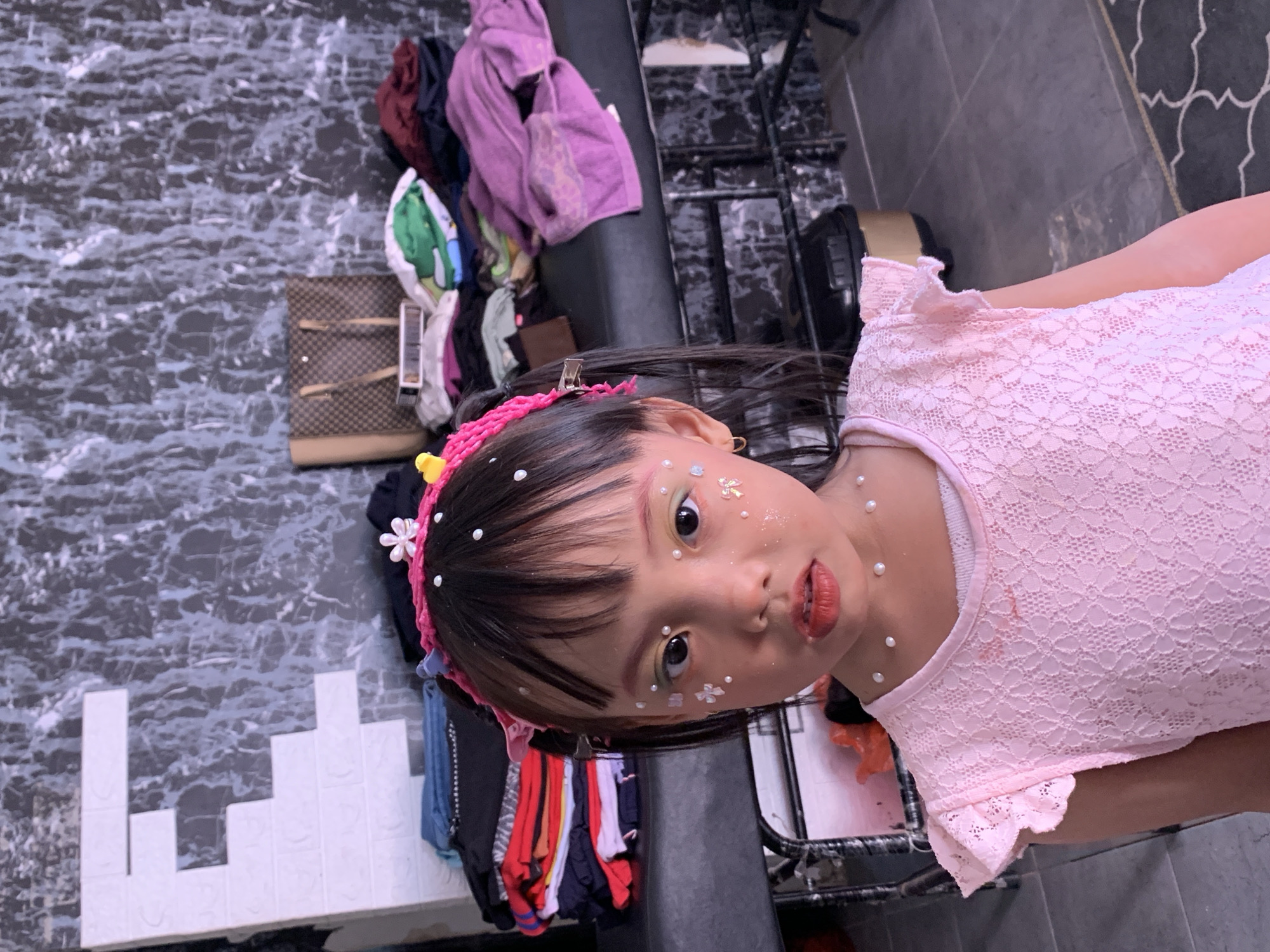 In a residential setting in Malang, Indonesia, a young girl is pictured. She has dark hair styled in pigtails and is adorned with face makeup, including glitter, pearls, and pink blush, and wears a pink lace dress. The background features a wall with a dark marble-like pattern and a large white "E" shaped decoration. Behind her, a dark-colored surface holds stacks of folded clothing and a designer handbag, suggesting a domestic or storage area. The floor is tiled with dark gray tiles.