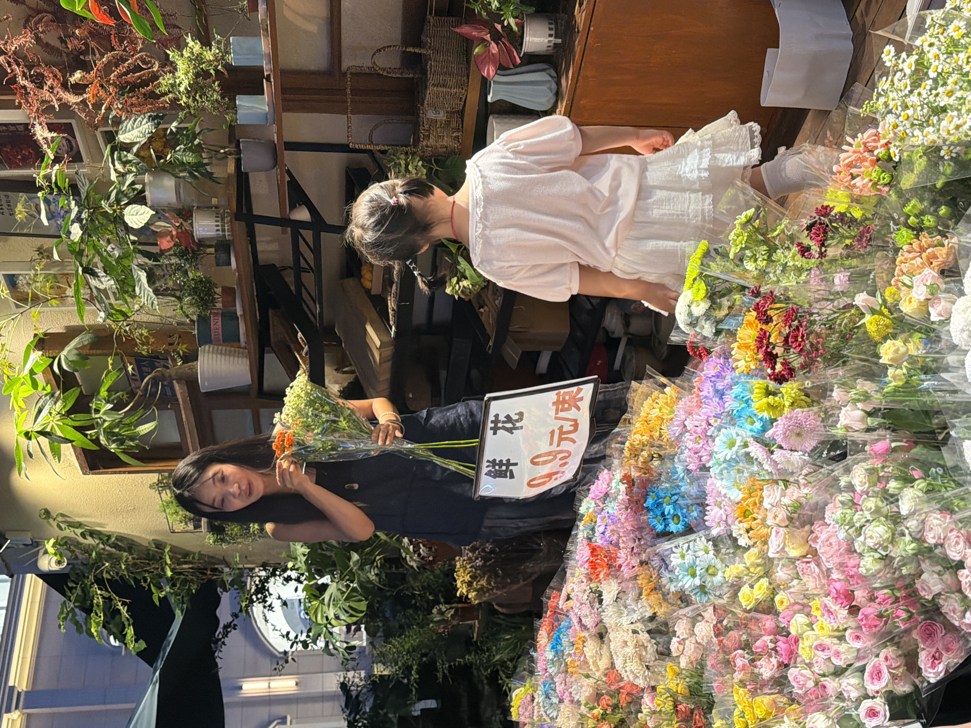 This media file captures a vibrant scene at a flower stand in Chongqing High-tech Zone, China. Two individuals, a young girl and an adult woman, are present. The girl, wearing a white dress with puffed sleeves, stands near a selection of potted plants. The woman, dressed in a dark top, is holding a bouquet of orange flowers and appears to be interacting with them.
The setting is an outdoor market or shop with a focus on flowers and plants. Various bouquets of brightly colored flowers, including roses and daisies, are prominently displayed, many wrapped in clear plastic. Potted plants are arranged on shelves and in the background, adding to the lush, botanical atmosphere.
A sign with Chinese characters is visible, indicating prices or descriptions of the flowers. The overall mood is cheerful and bright, with the sunlight suggesting it is daytime. The abundance of flowers and the presence of people engaging with them implies a lively commercial activity, likely flower sales. The image is oriented vertically.