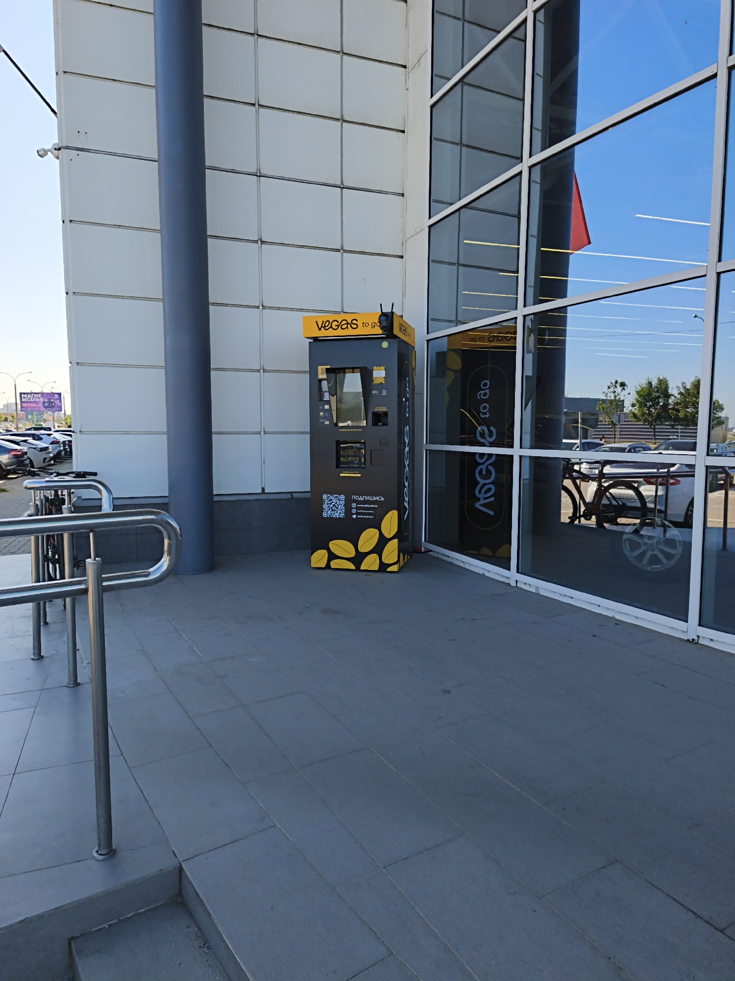 The image shows an outdoor scene in front of a modern building with a glass facade. A vending machine with the branding "VEGAS to go" is positioned to the left of the entrance. The machine is black and yellow and displays various icons and text in Cyrillic script, including "ПОДПИШИСЬ" (subscribe) and QR codes. To the left of the vending machine, there are chrome handrails and bicycle racks. The building's exterior is a combination of light-colored panels and large glass windows, which reflect the blue sky and some trees. In the background, cars are parked in a lot, and a billboard with Cyrillic text is visible. The ground is paved with large, grey tiles. The scene appears to be a public space, possibly near a shopping center or commercial area. Based on the Cyrillic script and context clues such as the billboard advertising "МАГИЯ МЕСЕЛЬ" (Magic of Mesel), this location is likely in Minsk, Belarus.