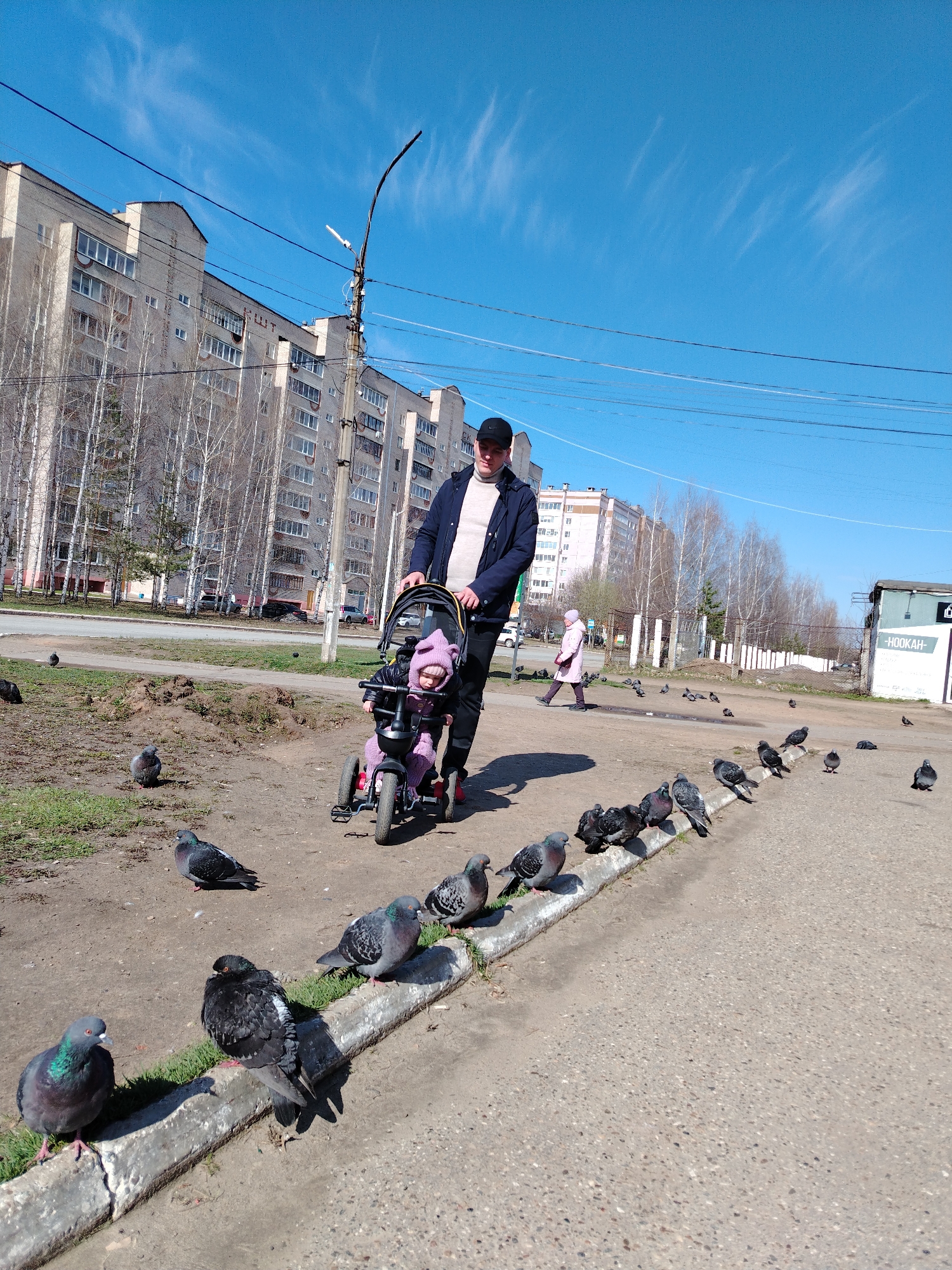 The image shows a man pushing a child on a tricycle in a public area in the city of Chaikovsky, Russia. The man is wearing a dark jacket, light-colored sweater, dark pants, and a black baseball cap. The child is wearing a purple outfit with a hat, riding a purple tricycle. There are numerous pigeons scattered on the ground and along the curb of the sidewalk. In the background, there are multi-story apartment buildings and bare trees, indicating a possible early spring or late autumn season. Other individuals can be seen walking in the distance. A utility pole is visible, along with overhead power lines, and a building with the word "HOOKAH" is on the right side of the image. The sky is bright blue with a few wispy clouds.