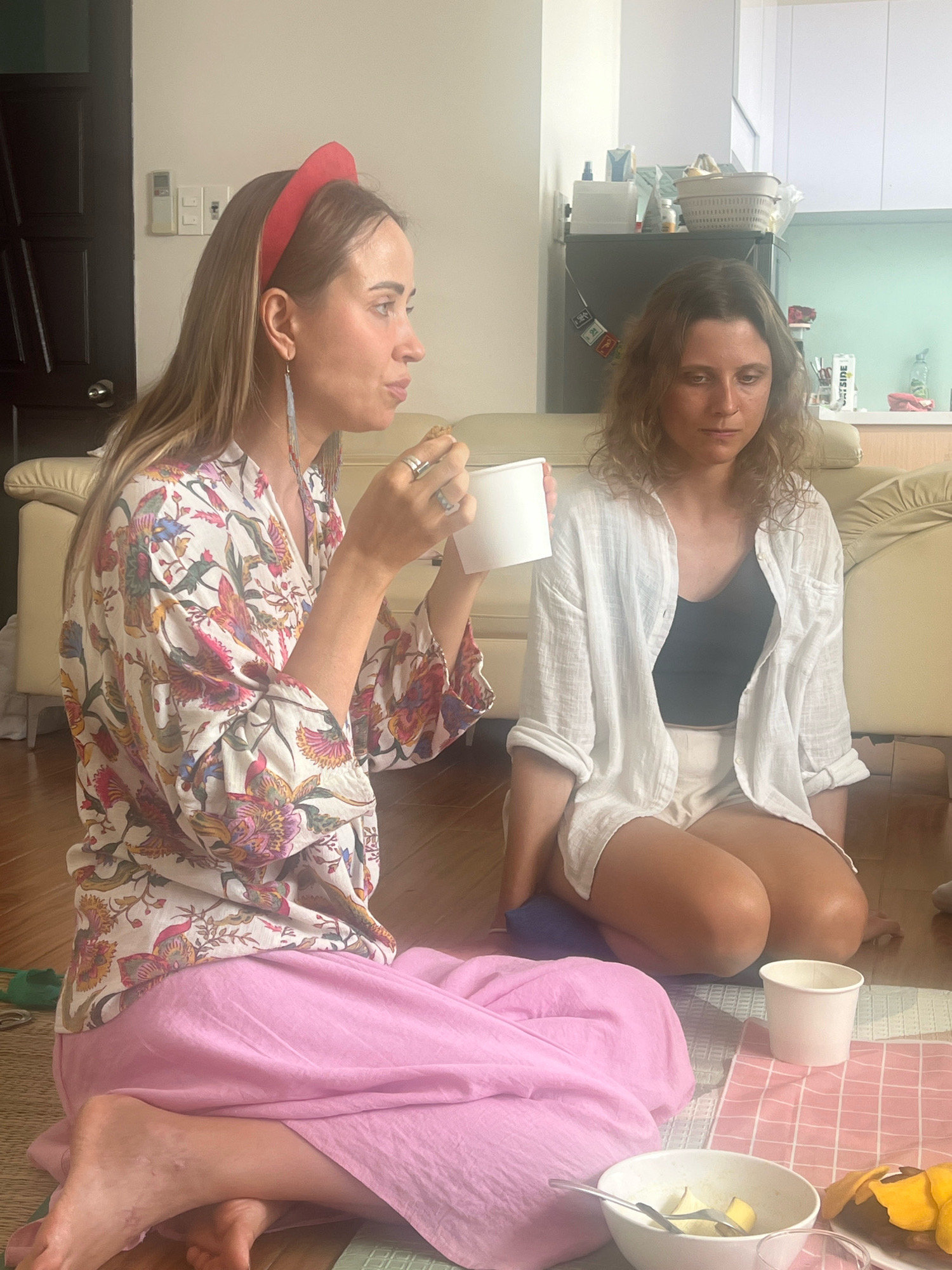 Two women are sitting on the floor in what appears to be a residential living room in Đà Nẵng, Vietnam. The woman on the left, with blonde hair, is wearing a floral print robe and a pink skirt. She is holding a white paper cup and appears to be eating something. She has a red headband on and long, dangling earrings. The woman on the right has brown, curly hair and is wearing a white open button-down shirt over a black tank top and white shorts. She is looking down with a pensive expression. They are sitting on a mat with a pink checkered pattern. In the foreground, there is a bowl of fruit and a plate of yellow food. Behind them, a couch and a kitchen area are visible, including a refrigerator and cabinets. The room is brightly lit, suggesting it is daytime.