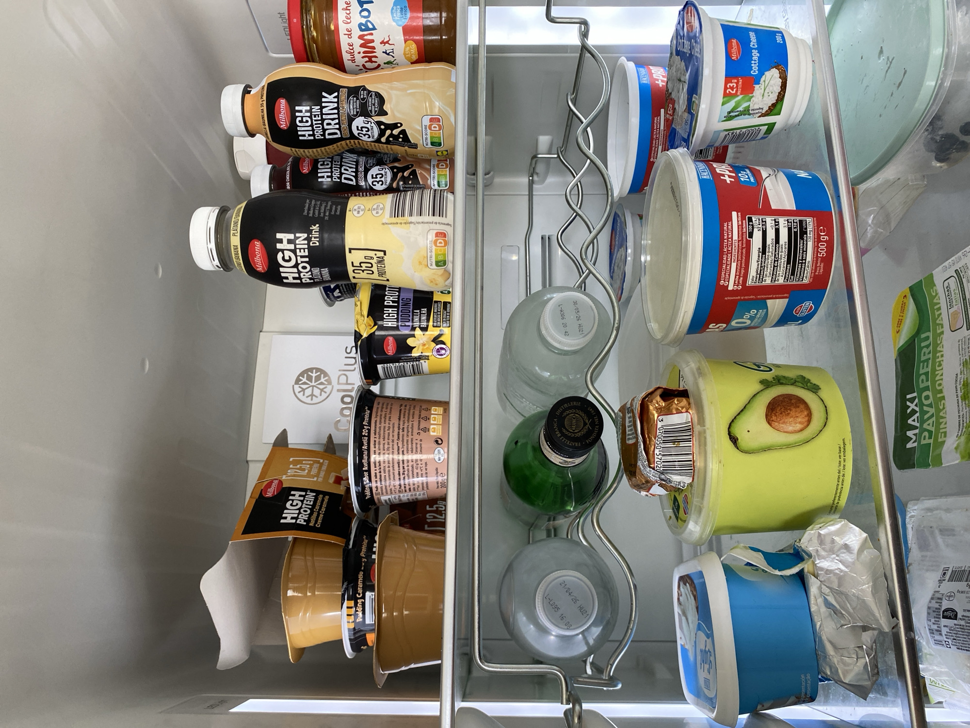 The image displays the interior of a brightly lit refrigerator, filled with various food and drink products. The perspective is looking down into the fridge, which is well-stocked and organized on several shelves and a bottle rack.
On the left side, a row of "Milbona High Protein Drink" bottles is visible. There are three chocolate-flavored bottles and one vanilla-flavored, each stating "35g PROTEINA" and displaying a Nutri-Score. Below these are several "Milbona High Protein" puddings in small plastic cups, including caramel (12.5g protein), vanilla, and hazelnut (20g protein) flavors. A white box labeled "CoolPlus" is also present.
The right side of the fridge contains a variety of dairy products and other items. On the top shelf, a jar of "dulce de leche CHIMBOT" is visible. Next to it are two "Milbona Cottage Cheese" containers, each stating "200g" and "23g PROTEINA". Several large plastic tubs, some white and blue, one with a red label indicating "0% GRASA" and "10g Proteínas" per 100g, likely contain natural yogurt or a similar dairy product.
A metal, wavy rack holds several bottles. A green glass bottle with "FRATELLI BRANCA DISTILLERIE" on the cap and two grey bottles with visible dates "24/04/26" and "30 05 26" are seen here. On the lowest visible shelf, a light green tub featuring an avocado image is present, alongside a blue tub partially covered in foil. A small, foil-wrapped item, possibly a chocolate bar, is placed on top of the avocado tub. Finally, a package of "MAXI PAVO PERU FINAS LONCHAS" (turkey breast slices) is also visible.
The packaging includes text in Spanish and Portuguese, suggesting a location where these languages are spoken, consistent with Gijón/Xixón, Spain. The scene depicts a typical, well-used refrigerator interior, indicative of daily food storage.