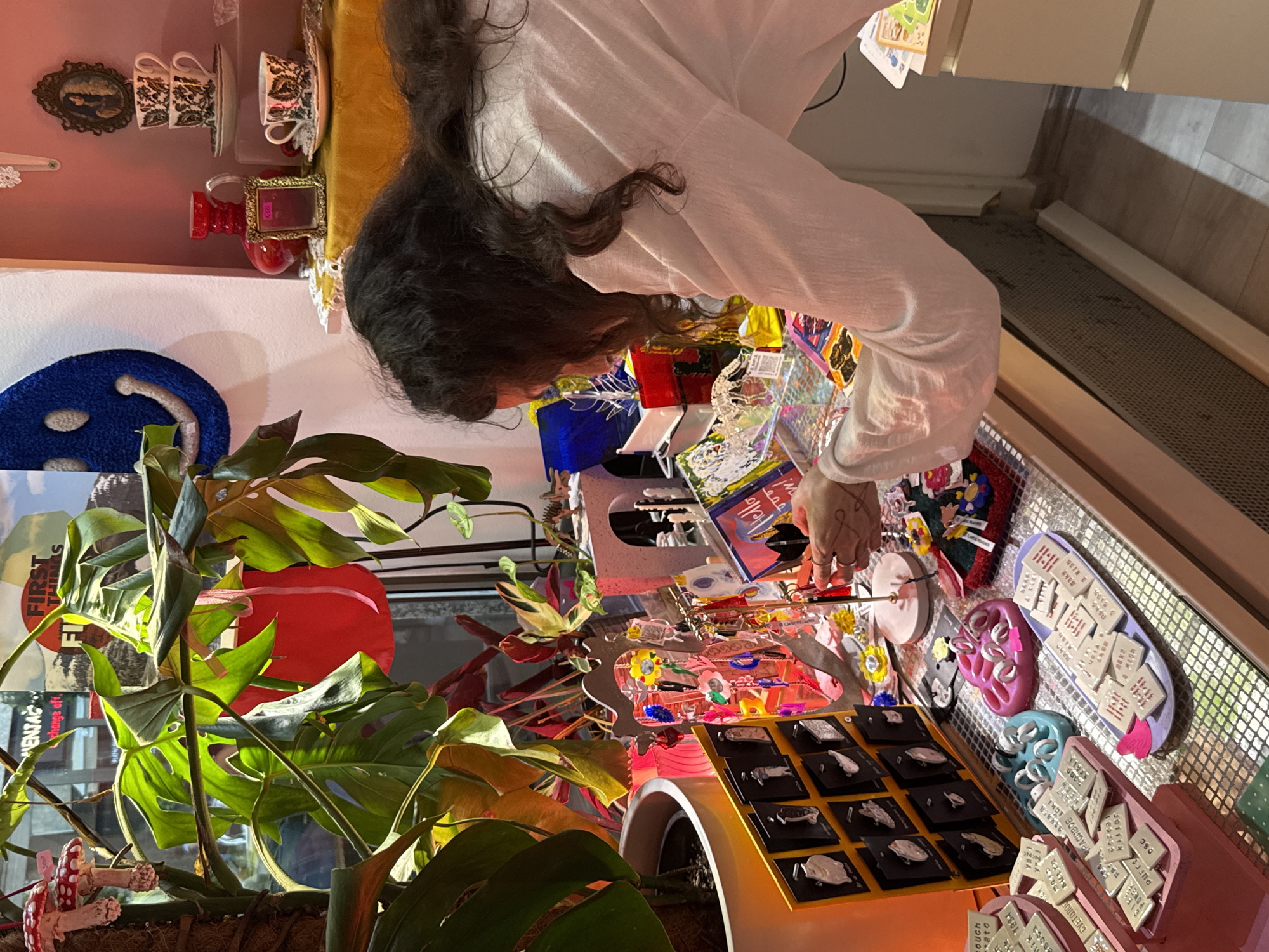 The image depicts a woman with dark, curly hair examining items on a display shelf in a brightly lit shop. She is wearing a light-colored, long-sleeved top. The display shelf is filled with various trinkets, including jewelry, charms, and small decorative items. In the foreground, there are trays of small, square-shaped objects with text, as well as pink and blue silicone molds holding rings. Behind her, a large green plant with broad leaves is visible, and further back, there are stacked teacups on a shelf, a red vase, and a blue smiley-face decoration. The shop appears to be filled with eclectic and colorful merchandise. The location is Vracar Urban Municipality, Serbia.