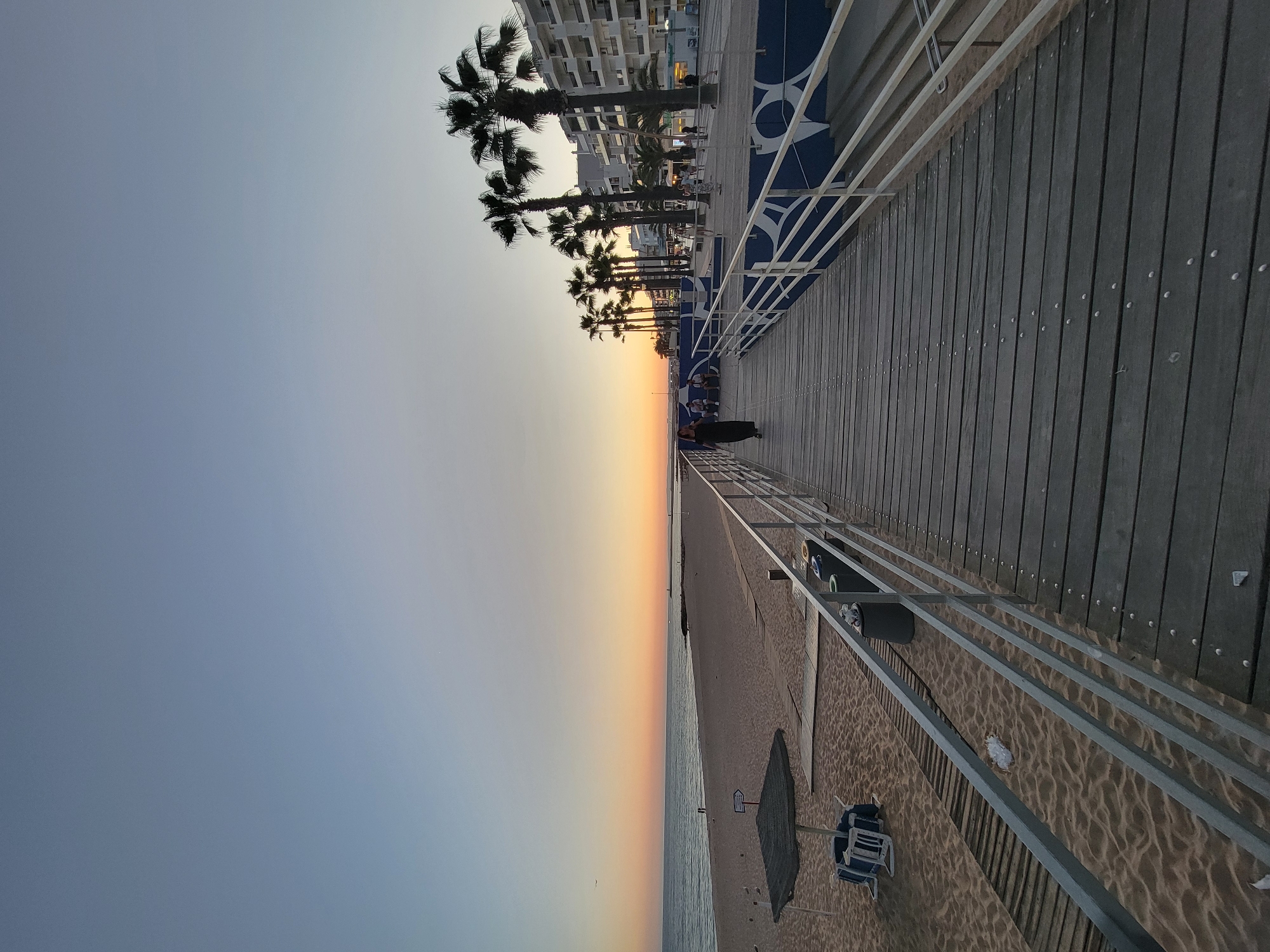 The image depicts a sunset view from a wooden boardwalk along a sandy beach in Quarteira, Portugal. The sky transitions from a pale blue at the top to warm hues of orange and yellow near the horizon. The ocean is calm and stretches to the left of the frame, meeting the sandy beach. On the beach, there are a few beach chairs and an umbrella. To the right, the boardwalk extends into the distance, bordered by white railings. Further to the right, several palm trees stand in front of a row of multi-story buildings. A few people are visible walking on the boardwalk and sitting on benches along the promenade. One person in a dark dress is walking towards the camera on the boardwalk, and several other individuals are in the distance on the promenade. The overall scene suggests a peaceful evening at a coastal resort town.