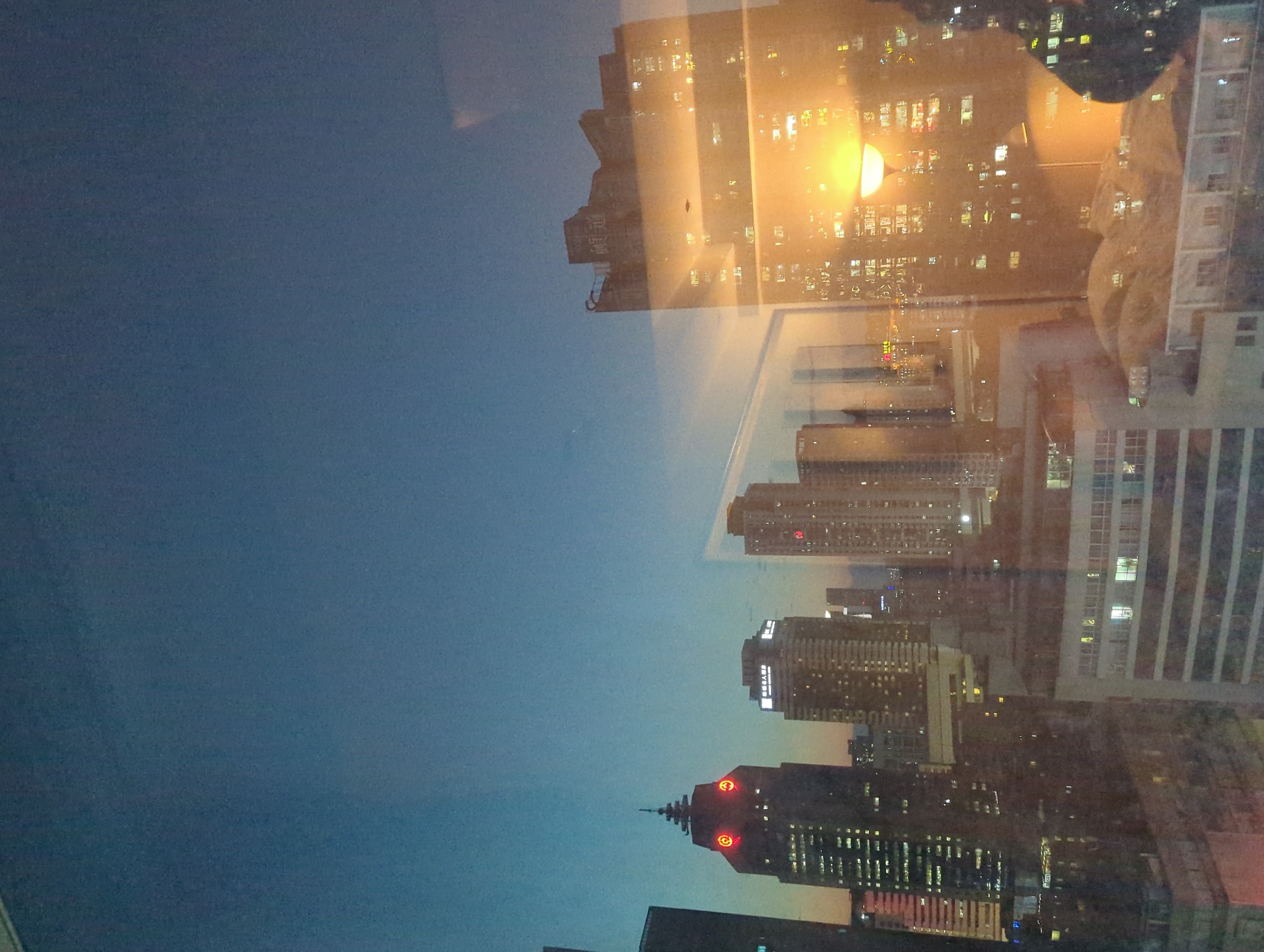 The image depicts a twilight cityscape with various buildings, some of which are illuminated by artificial lights. A prominent building on the left has a distinctive red logo or symbol resembling a stylized 'Q' or a face with glowing eyes. To the right, a reflection is visible in a glass surface, showing a building with illuminated windows and a bright, glowing light fixture that appears to be a streetlamp. The sky transitions from a deep blue at the top to a lighter, warmer hue near the horizon. The overall impression is of an urban environment at dusk, with the city lights beginning to dominate the darkening sky. The image's composition suggests it was taken from an elevated vantage point, possibly a window. The specific location of Qingyang District, China, is mentioned in the prompt but is not visually identifiable from the provided image.