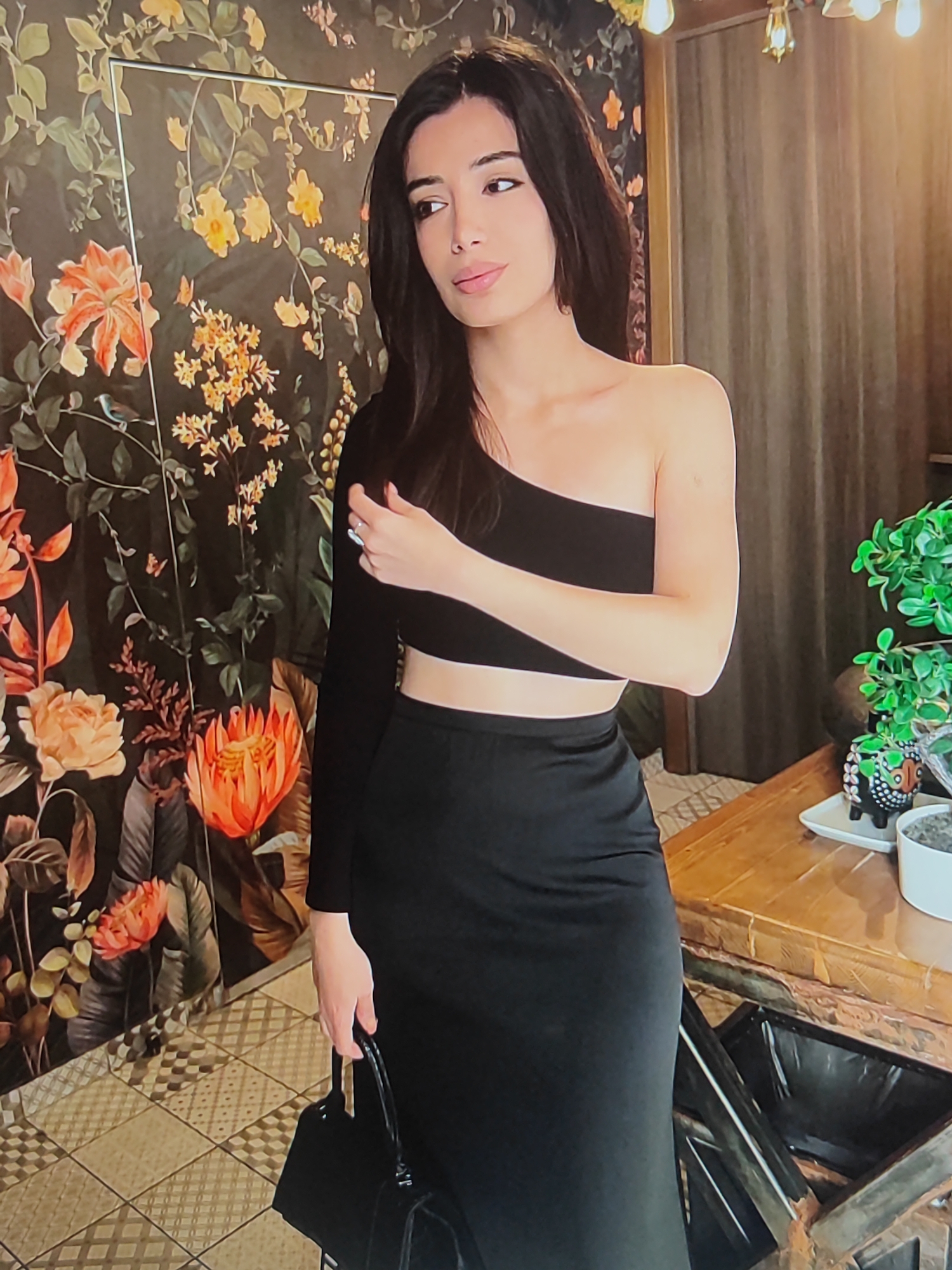 A young woman with long, dark hair stands in front of a floral wallpaper. She is wearing a black, one-shoulder crop top and a black, floor-length skirt. She is holding a black purse in her right hand. In the background, there are hanging lights and some potted plants on a wooden table. The wallpaper features large orange and yellow flowers with green leaves. The floor is tiled with a patterned design. The lighting suggests it is indoors, possibly during the day or evening. The woman appears to be looking off to the side with a neutral expression.