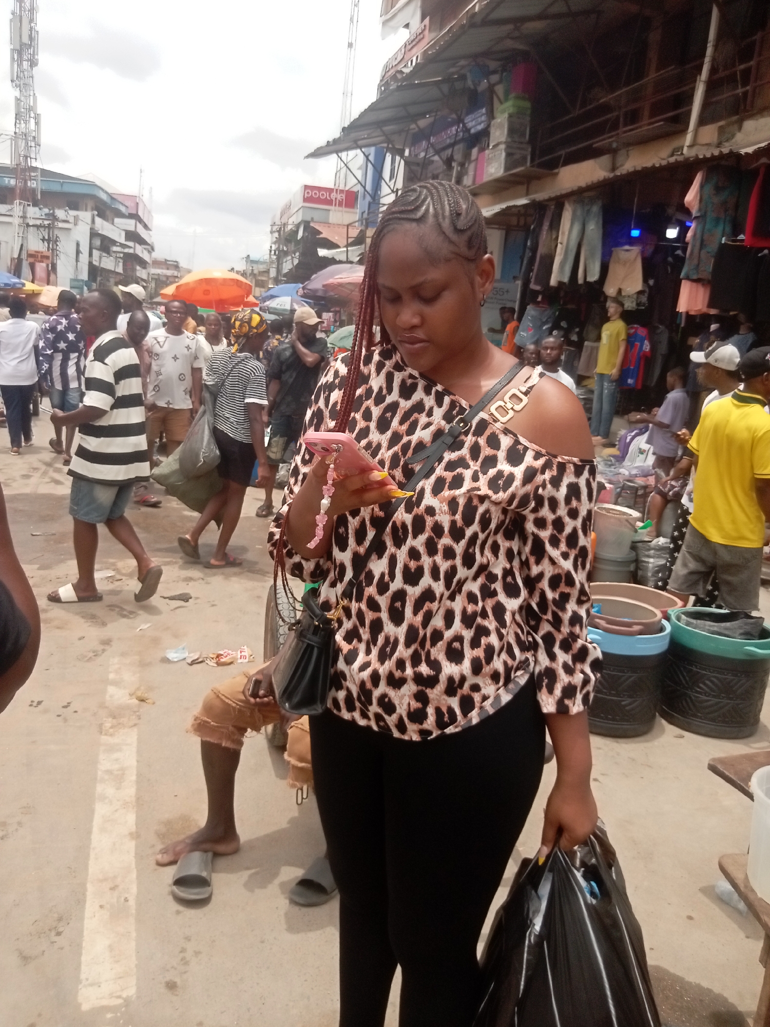 The image depicts a woman in a crowded outdoor market scene in Fegge, Nigeria. The woman is wearing a brown and black leopard-print blouse, black pants, and has braided hair. She is holding a black shopping bag in her right hand and looking at her pink smartphone in her left. She has a black purse hanging over her shoulder.
The background reveals a busy market environment with several people walking around. Some are wearing striped shirts, and others are wearing casual clothing. There are vendor stalls with items for sale, including clothes. Orange umbrellas are visible in the distance.
The ground appears to be concrete and littered with debris. A man seated on the ground with worn clothing is partially visible to the left. There are several buckets behind the woman to the right.
