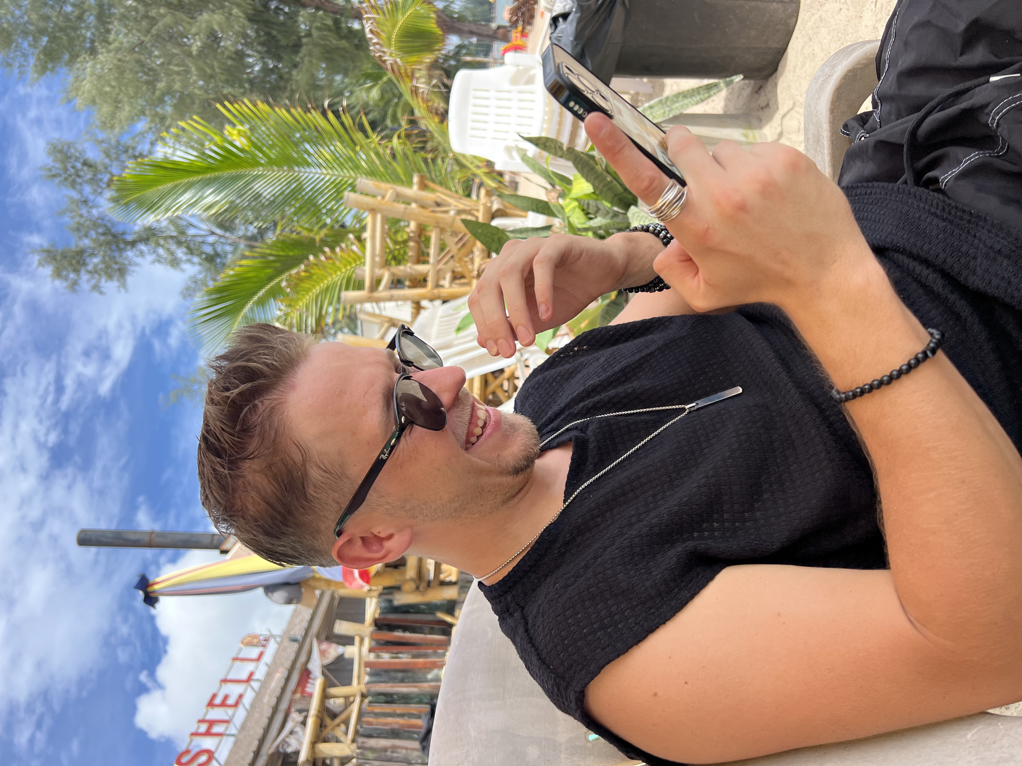A man with blond hair and sunglasses sits in a chair outdoors, looking at his phone and smiling. He is wearing a black sleeveless shirt and a necklace. He has bracelets on his wrist and rings on his fingers. In the background, there are palm trees, a building with a sign that says "SHELLA," and plastic chairs. The sky is blue with clouds.