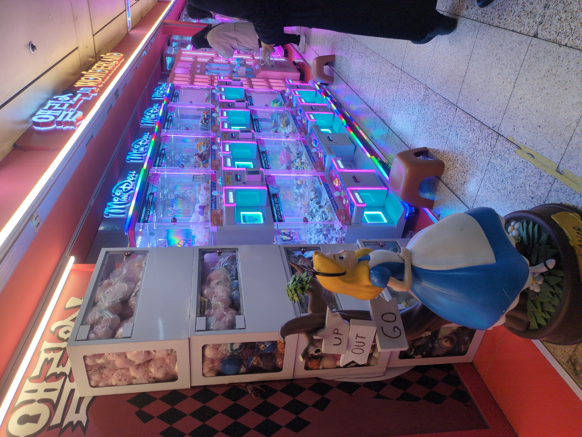 Here's a detailed description of the image:
**Location:** The image appears to be taken inside an arcade, likely located in Seoul, South Korea, indicated by Korean signage.
**Environment:** The arcade features a long row of claw machine games. These machines are illuminated with bright neon lighting, mostly in pink, blue, and green. The flooring has a mosaic-like pattern.
**Objects:**
* **Claw Machines:** Numerous claw machines are visible, each filled with plush toys or other prizes.
* **Seating:** A small, brown stool is present near the claw machines.
* **"Alice in Wonderland" Statue:** An "Alice in Wonderland" statue stands near the edge of the frame. The statue has signs.
* **Signage:** There are neon signs in Korean and English ("Wonderland", "Catch Star") above the machines. The signs say "UP, OUT, GO."
**People:**
* The image shows what appears to be the legs of some people standing behind the machines.
**Actions/Interactions:**
* It appears that people are actively engaging with or observing the claw machines.