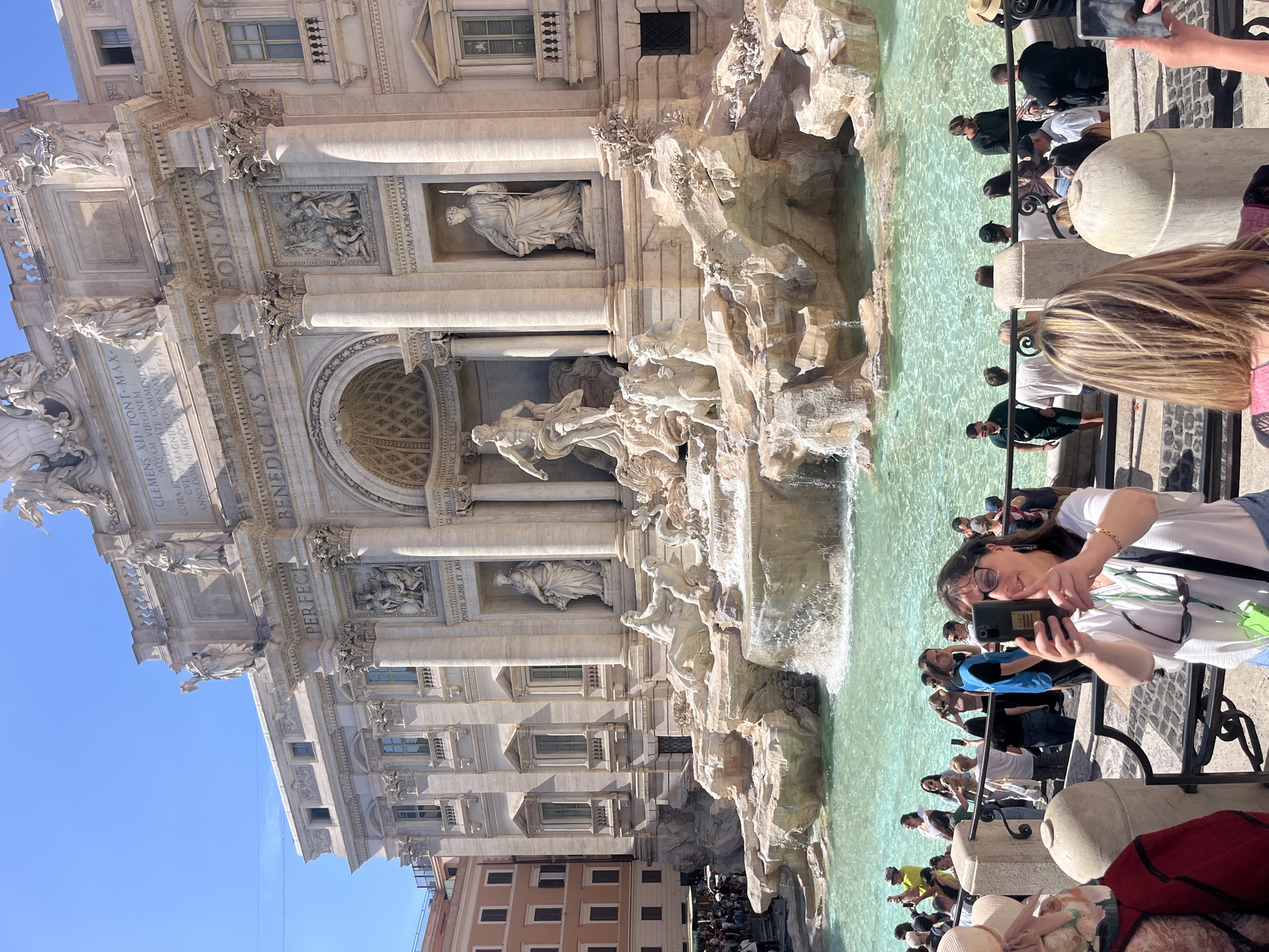 This media file captures a vibrant scene at the iconic Trevi Fountain in Rome, Italy, on a bright, sunny day. The image is composed with the grand Baroque fountain dominating the left side, featuring its intricate sculptures, grottoes, and flowing turquoise water. The central sculpture depicts Oceanus in a shell-shaped chariot pulled by sea horses and tritons, flanked by allegorical statues. Above the fountain, the facade of Palazzo Poli rises with classical columns, cornices, and windows.
A crowd of tourists gathers around the fountain's edge, visible on the right side of the image. Many are actively engaged in sightseeing, taking photos, and admiring the monument. In the foreground, a woman wearing glasses and a white shirt is prominently featured, holding a smartphone to take a selfie or photograph the fountain. Her expression appears pleasant. Other individuals are seen from behind or partially obscured, leaning against the protective railing that separates the viewing area from the water. Hats, bags, and various casual clothing indicate a warm weather setting. The sky is a clear, brilliant blue, suggesting midday or early afternoon sunshine.
Visible Latin inscriptions on the fountain's facade, confirmed by the OCR, include:
"CLEMENS XII PONT MAX AQVAM VIRGINEM COPIA ET SALYBRITATE COMMENDATAM CVLTV MAGNIFICO ORNAVIT ANNO DOMINI MDCCXXXV PONTIF-VI" (Pope Clement XII renovated the Aqua Virgo aqueduct, recommending it for abundance and health, magnificently adorning it in the year of the Lord 1735, sixth year of his pontificate).
"BENEDICTUS XIV PON MA PERFECI" (Benedict XIV, Supreme Pontiff, completed it).
"SOLVTVM A DOM MDCCLXII POSITIS SIGNIS ET AN GLYPHIS TANLIS IVSSY CLEMENTIS XI PONTE" (Dedicated in the year of the Lord 1762, with the placement of the statues and carvings by order of Clement XI).
The image conveys a bustling atmosphere typical of a popular tourist destination.