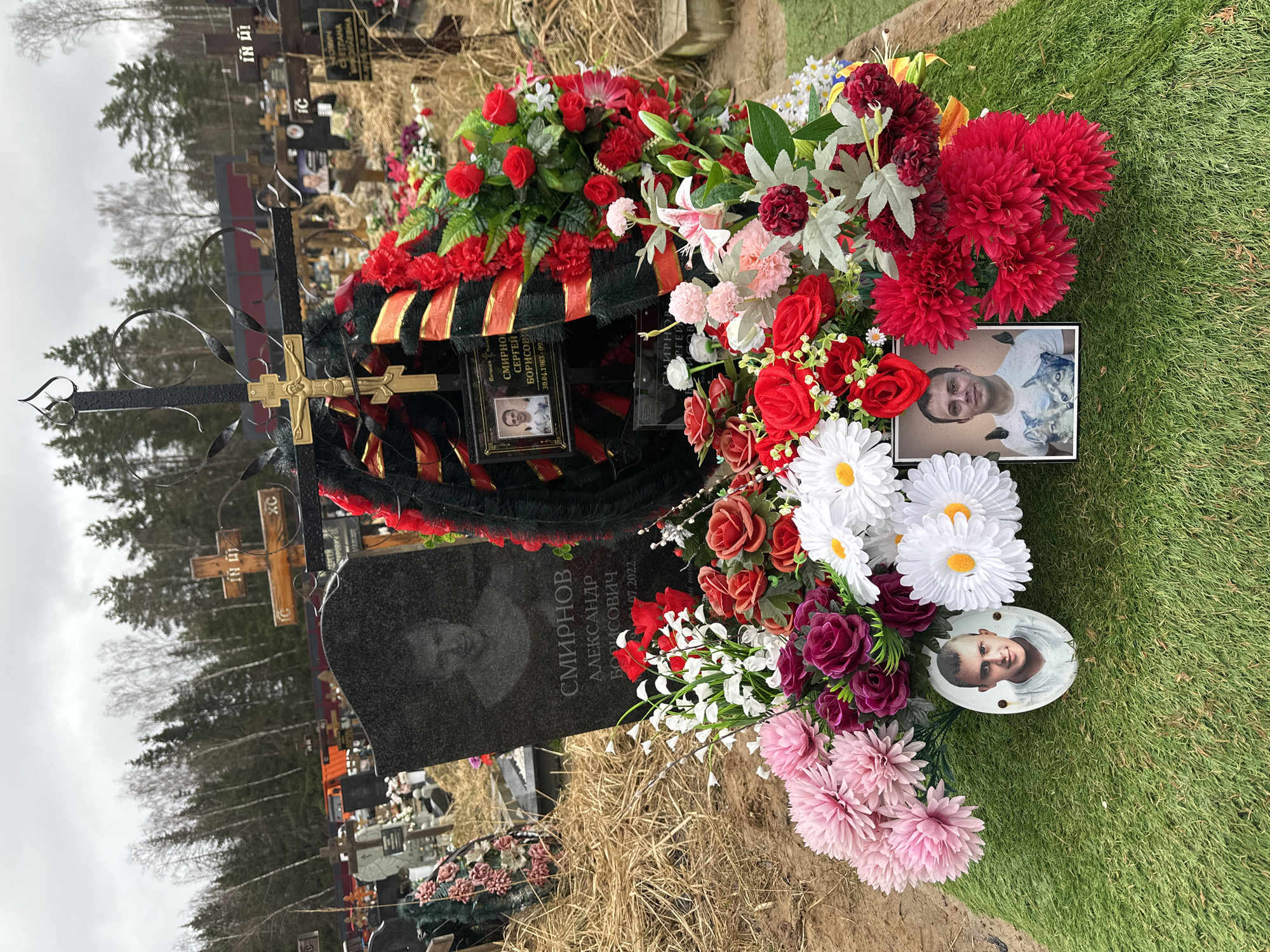 Here is a description of the image:
The image shows a gravesite, presumably located in a cemetery. The grave has a black headstone with a photograph of a man etched into it and a name written in Cyrillic: "СМИРНОВ АЛЕКСАНДР БОРИСОВИЧ", alongside what is presumably the date of death: "07.2022". There is a wrought iron cross behind the headstone.
The grave is adorned with numerous floral arrangements, including a wreath of dark green and red artificial flowers with gold and black ribbon. There are also fresh and artificial flowers in a variety of colors including red, white, pink, and burgundy. There are two framed photos and one ceramic photo of the deceased placed among the flowers. One frame reads "СМИРНОВ СЕРГЕЙ БОРИСОВ" and presumably includes the date of birth and death.
The ground around the grave is covered in grass. Other graves and trees are visible in the background. The sky appears overcast.
I could not independently verify that the image is from Шарапово, Russia.