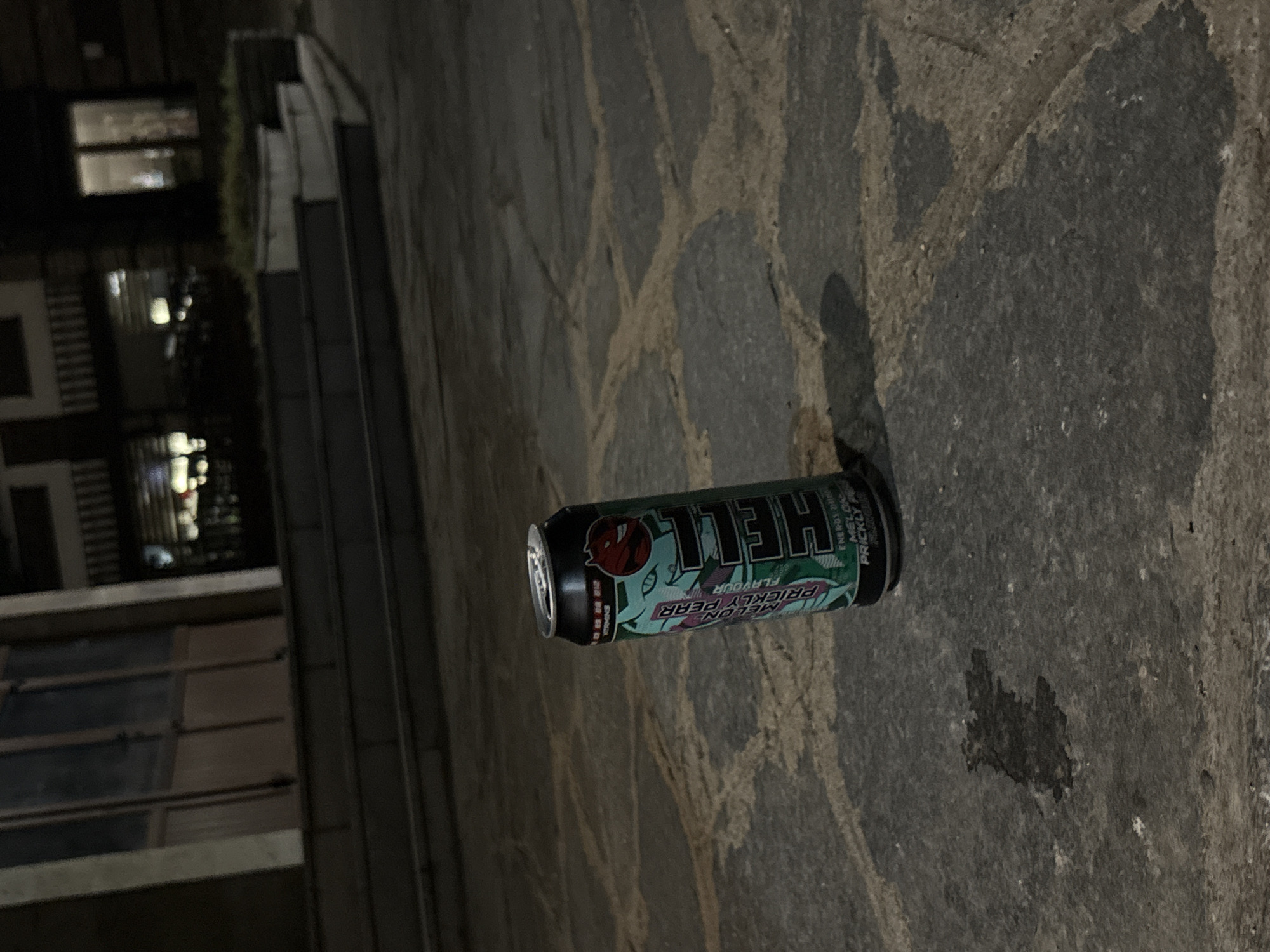 A single, partially consumed can of "Hell" energy drink, specifically "Melon Prickly Pear" flavor, rests upright on a stone-paved sidewalk at night in the city of Sopot, Bulgaria. The can is predominantly black and mint green, featuring a cartoon devil logo and text indicating "ENERGY DRINK" and the flavor. The pavement consists of irregularly shaped, grey and tan stones. In the background, a set of concrete steps leads up to a building with large windows and doors, which appear to be dimly lit from within. The overall scene suggests an urban outdoor setting after dark, with no people present.