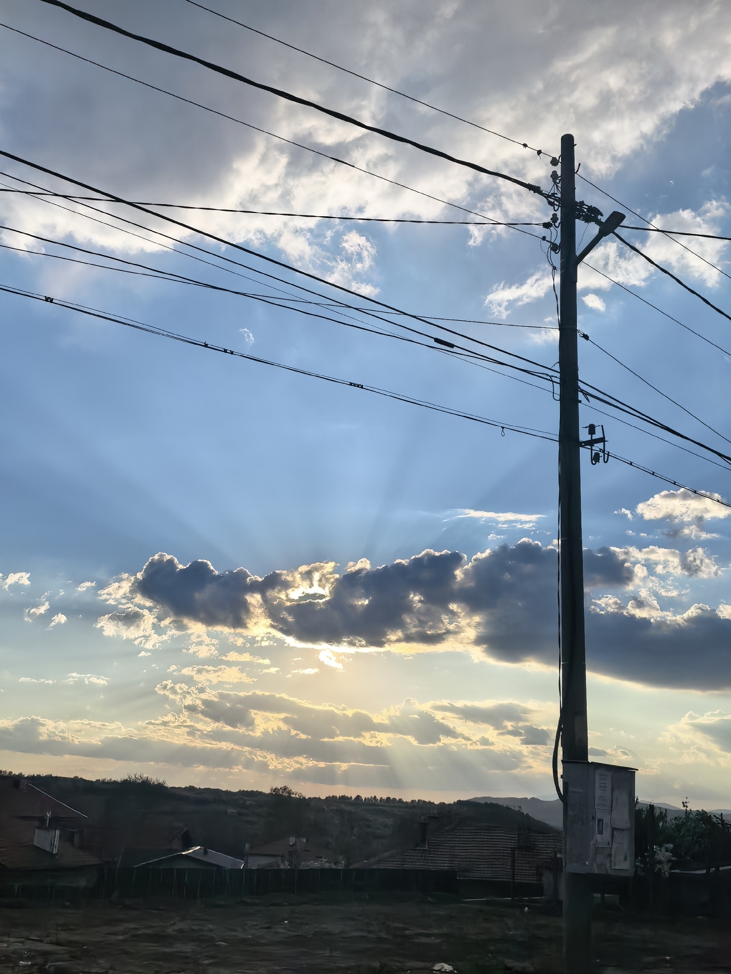 A vertical outdoor photograph captures a dramatic sky at what appears to be late afternoon or early evening, likely during sunset or sunrise. The upper two-thirds of the image are dominated by a vibrant blue sky interspersed with scattered cumulus clouds. Prominent sunrays, also known as crepuscular rays, fan out from behind the clouds in the mid-sky, indicating the low position of the sun.
A tall, dark utility pole stands on the right side, extending from the lower part of the frame almost to the top. Numerous power lines crisscross the sky from various angles, creating a complex network of dark lines against the bright and cloudy backdrop. The pole is equipped with a street light fixture near the top and an electrical box attached near its base.
Below the horizon line, the landscape is largely silhouetted and appears dark. It consists of undulating hills or terrain with sparse vegetation, and the faint outlines of what seem to be building rooftops are visible on the left side, nestled among the dark contours of the land. The overall scene is atmospheric, characterized by the interplay of light and shadow, with the sun's rays serving as a notable detail. No discernible text is visible on any object in the image.