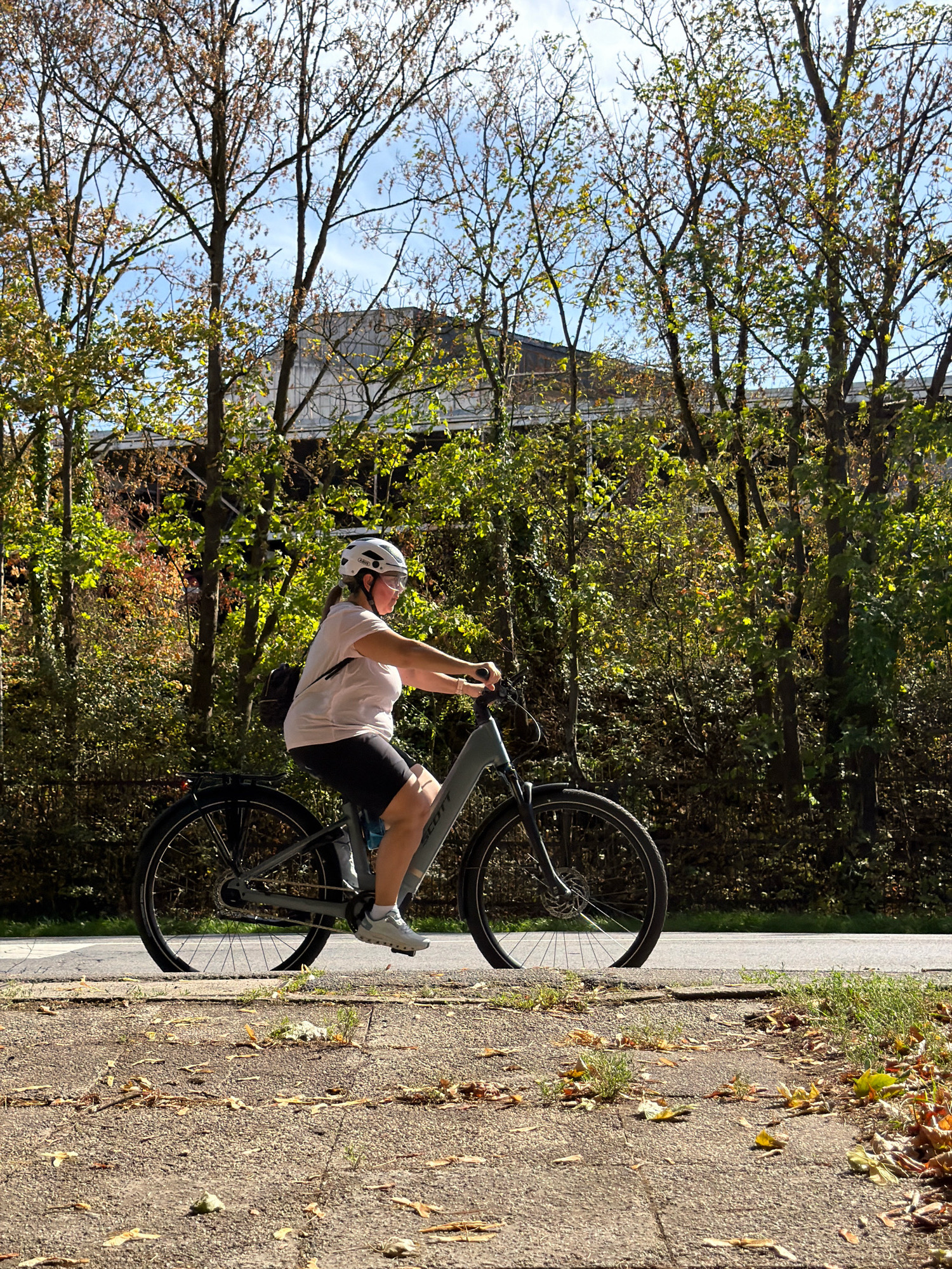 A woman is riding a grey electric bicycle on a paved path in Esch-sur-Alzette, Luxembourg. She is wearing a white helmet, clear glasses, a light pink t-shirt, and black cycling shorts. The bicycle has a backpack attached to the rear rack. The path is lined with trees displaying autumn foliage, with a building visible in the background. The ground in the foreground is a rough-textured paved surface scattered with fallen leaves. The sky is blue with some clouds.