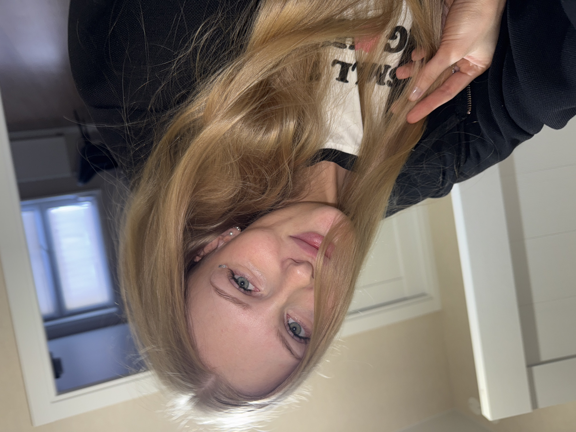 A young Caucasian woman with long blonde hair and blue eyes is taking a selfie in what appears to be a residential interior. She is wearing a black jacket over a white t-shirt with some text on it that is partially visible. Her hair is falling across her face, and she is holding a strand of it with her left hand. She has subtle makeup on, including eyeliner and possibly some glitter under her left eye. The background shows a doorway and part of a room with windows and a radiator. The lighting is soft, suggesting indoor lighting. There is no information in the image to indicate the specific location within the city of Perm, Russia.