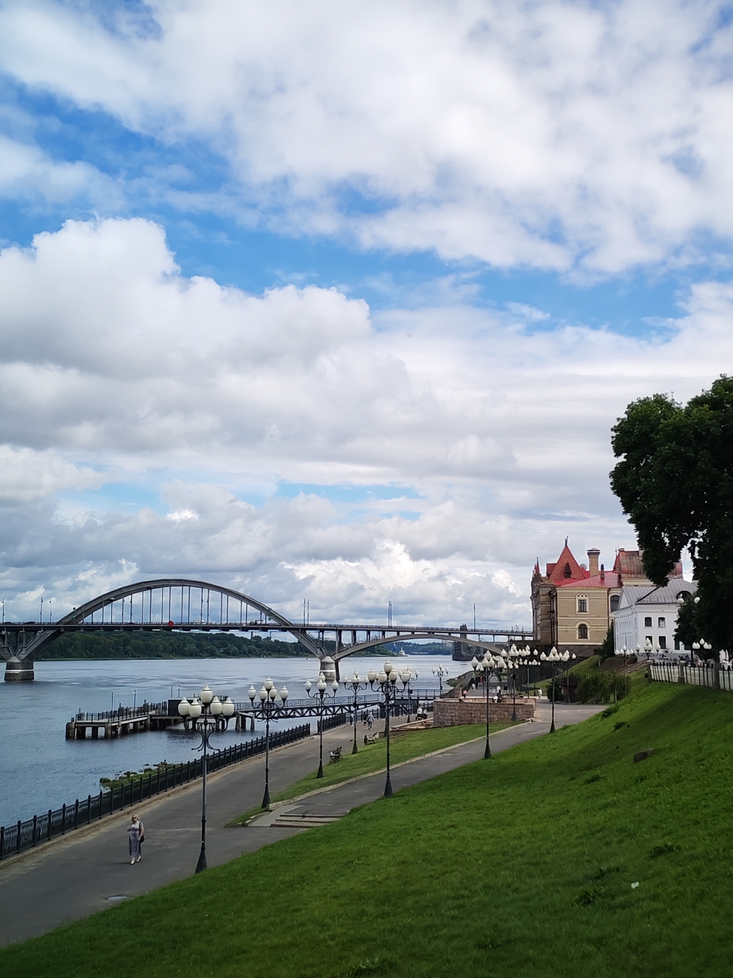 The image depicts a wide river with a large arched bridge spanning across it. On the right bank of the river, there is a paved promenade lined with ornate lampposts and lush green grass on a slope. Several buildings, including one with a distinctive red roof, are visible along the promenade. A lone woman in a light-colored dress is walking on the promenade. In the background, another bridge is visible further down the river, and a dense line of trees forms the far bank. The sky is partially cloudy with patches of blue. The scene is set in Bol'shie Mkhi, Russia, suggesting a tranquil urban waterfront.