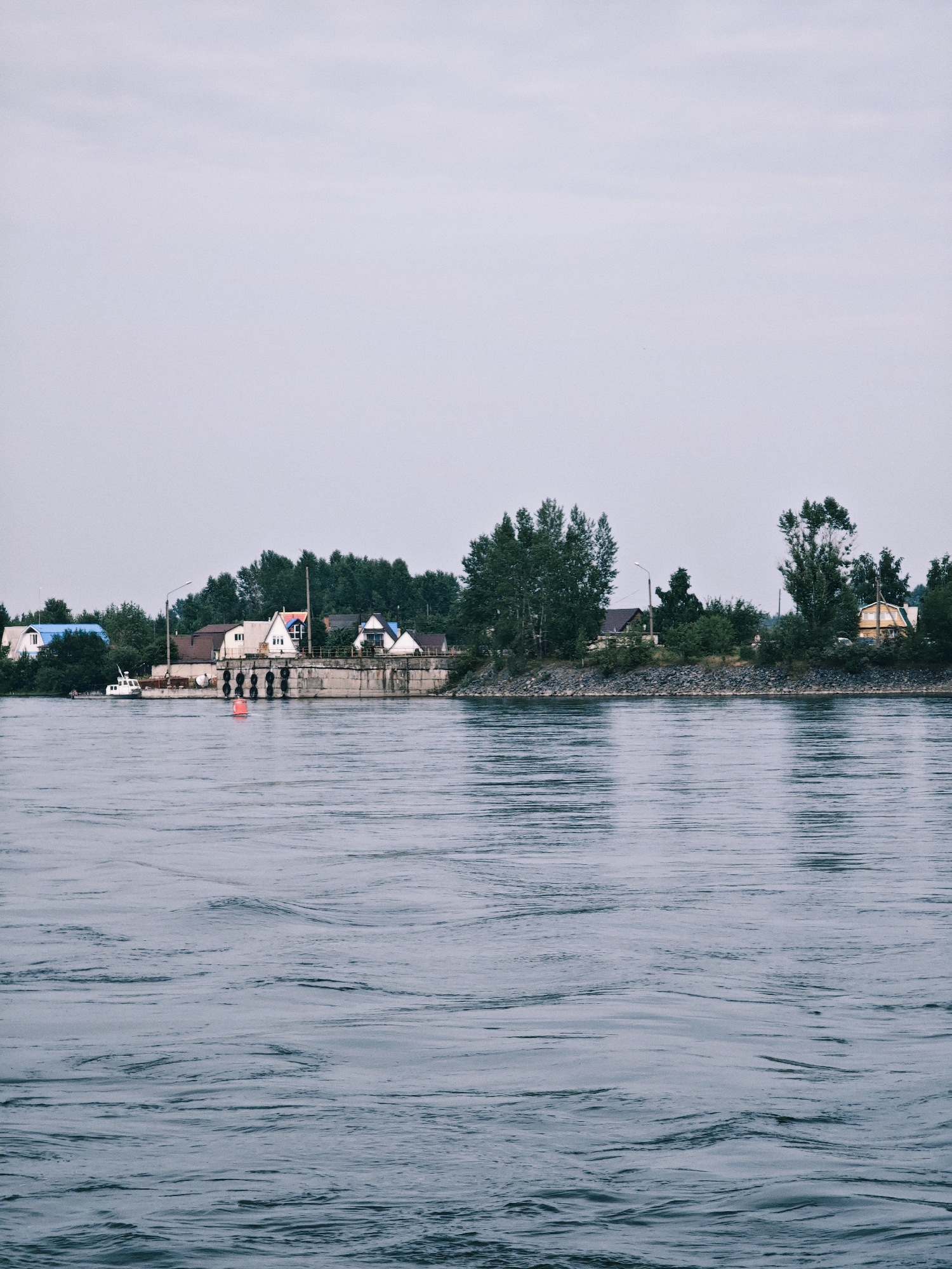 The image depicts a serene, overcast day by a body of water in Sosnovoborsk, Russia. The foreground is dominated by the rippling surface of a wide river, its water a deep, muted blue. In the middle ground, a concrete embankment lines the shore, punctuated by large, dark, circular fenders. A bright orange buoy floats in the water near the embankment.
Along the embankment, a small cluster of houses and buildings form a settlement. These structures vary in design, with some featuring pitched roofs in colors like blue and red. Lush green trees and foliage densely cover the area behind the houses, creating a natural barrier. A white boat is docked to the left of the embankment, and a street lamp stands tall near the buildings. The background is a pale, hazy sky, suggesting either dawn, dusk, or a heavily overcast day. The overall impression is one of quiet, rural life on the waterfront.