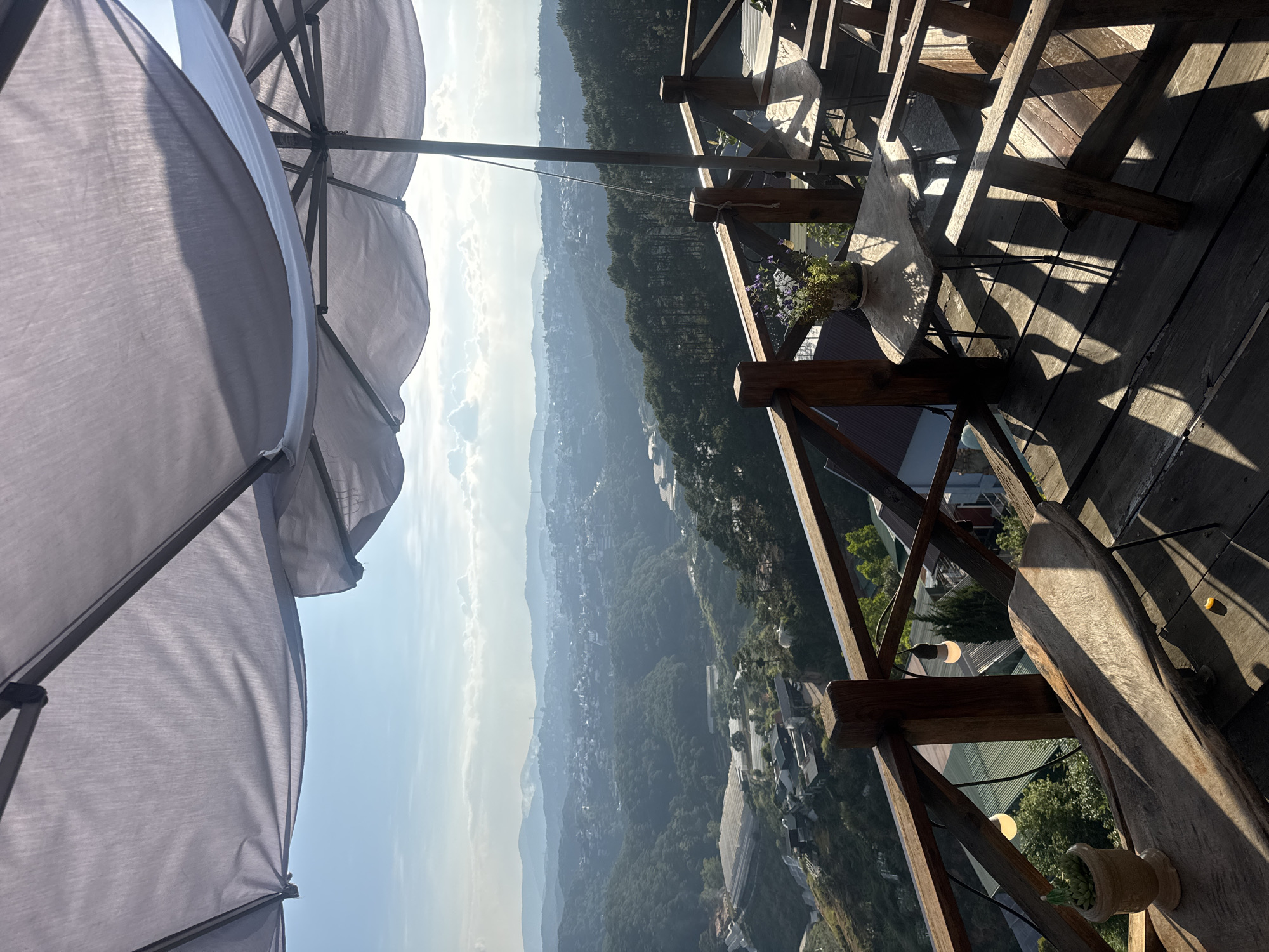 This image shows a view from a wooden balcony in Da Lat, Vietnam. The landscape is a mountainous area with trees and some buildings visible in the distance. In the foreground, there's a wooden railing and a section of a wooden table and chairs on the balcony. A flower pot with small purple flowers sits on the table. A large gray umbrella partially covers the top left of the frame. The lighting suggests it's daytime. The weather appears clear.