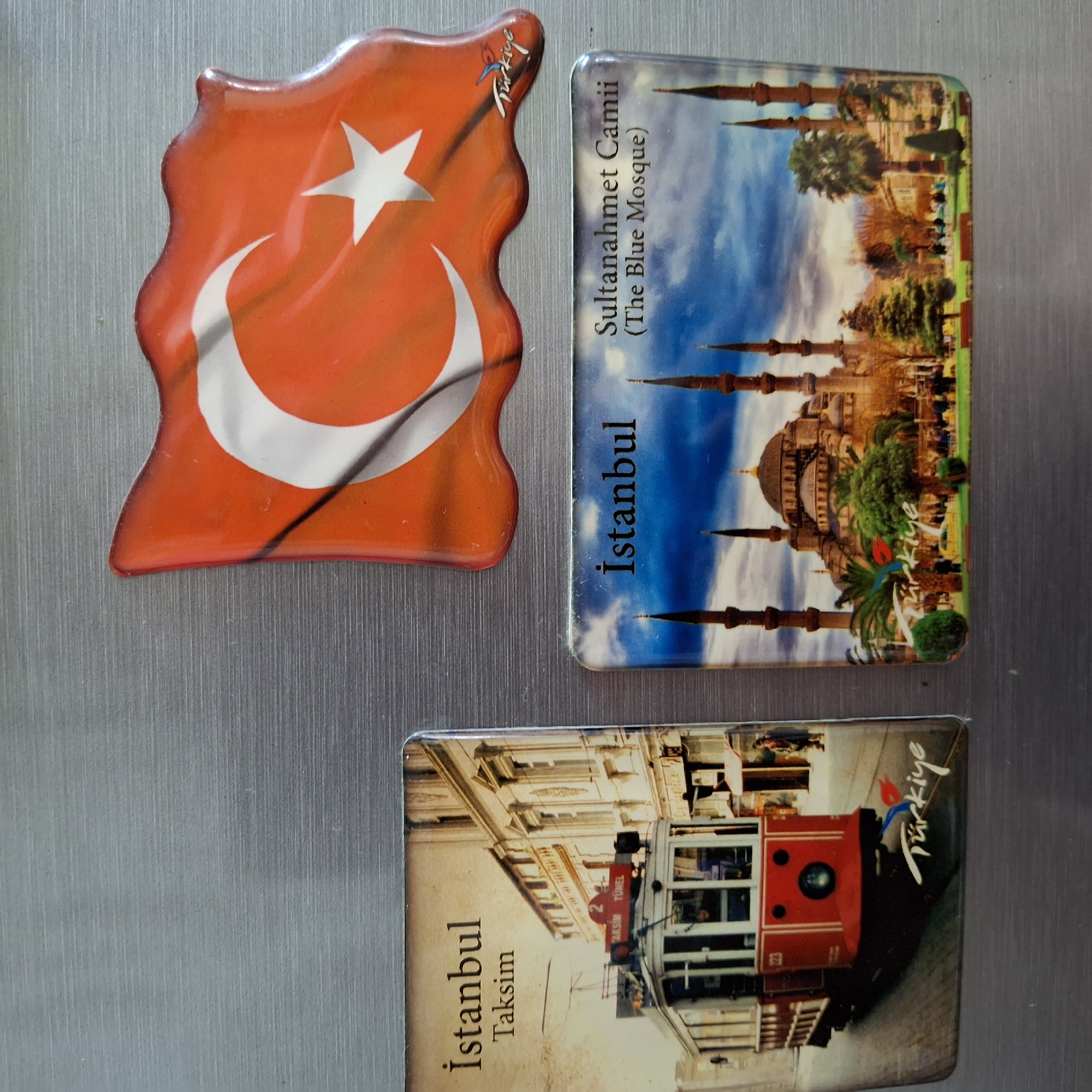The image displays three refrigerator magnets on a metallic surface. The leftmost magnet features a vintage red tram on a street with buildings in the background, and the text "Istanbul Taksim" and "Türkiye". The top right magnet is a Turkish flag in a wavy shape. The bottom right magnet shows a mosque with minarets, palm trees, and people in the foreground, with the text "Istanbul" and "Sultanahmet Camii (The Blue Mosque)" and "Türkiye".