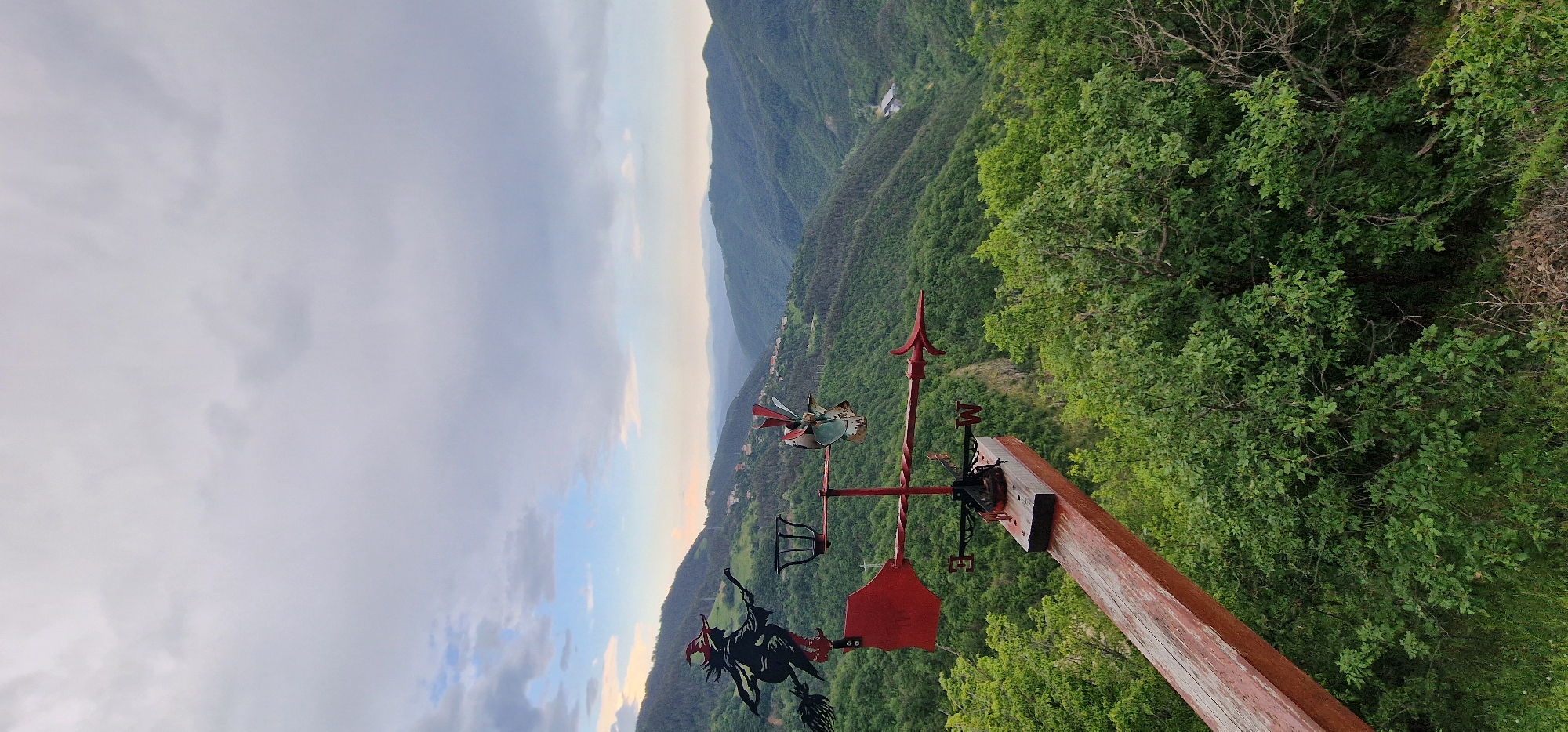 The image shows a landscape view, likely from a high vantage point in the Pechinska area of Bulgaria. A red and black weather vane is in the foreground, mounted on a wooden post, depicting a witch on a broom. Beyond the weather vane, a mountainous valley stretches into the distance under a partly cloudy sky. Lush green trees cover the lower slopes of the mountains. Some houses are visible in the valley below.