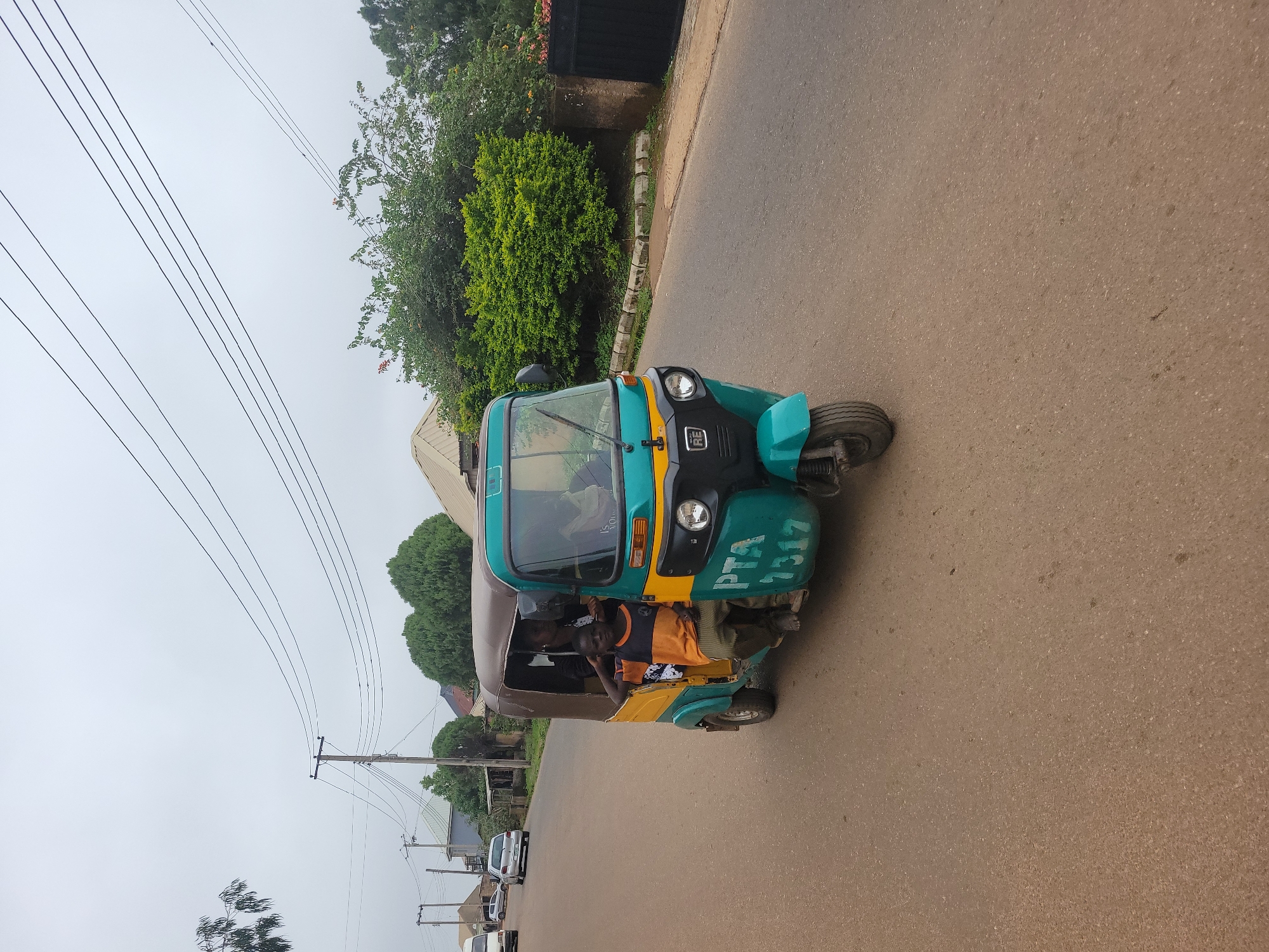 A teal and yellow auto-rickshaw, also known as a "Keke Napep," drives down a paved road in Jos, Nigeria, under an overcast sky. The auto-rickshaw has a brown canvas covering, and the front displays the letters "PTA" and the number "7317" in white. Inside the auto-rickshaw, a young boy with dark skin and short hair, wearing an orange and black striped t-shirt and green pants, sits in the front passenger seat with his left arm resting on his head. Behind him, a woman with dark skin and dark hair, wearing a dark top, is visible. The road is lined with lush green bushes and trees, and in the background, houses with corrugated iron roofs are visible. Power lines stretch across the sky, supported by wooden utility poles. A black metal gate stands to the right of the frame, partially obscured by foliage. Other vehicles, including a white van and a silver car, are seen in the distance on the road.