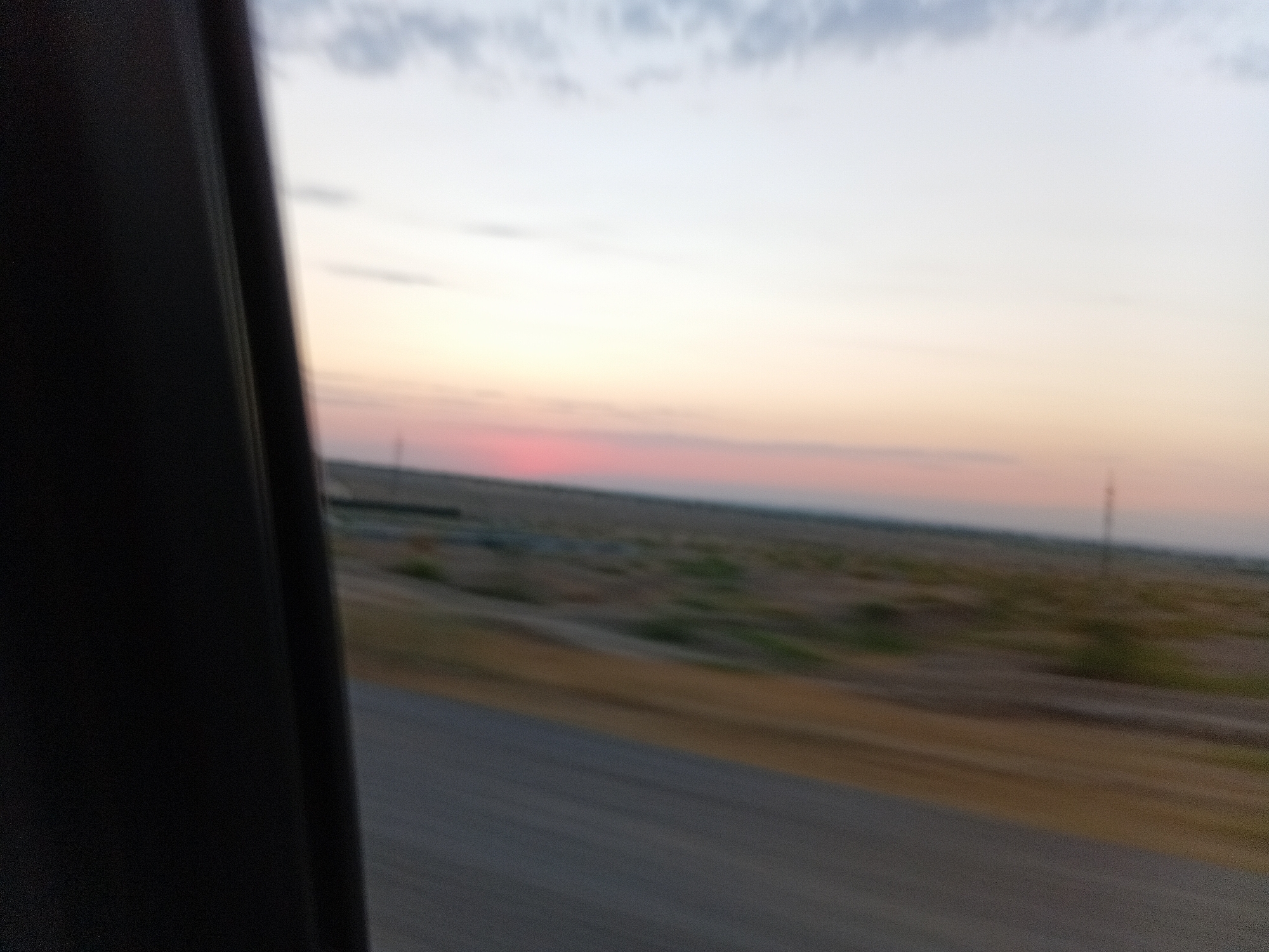 The image, captured from inside a moving vehicle, depicts a blurred desert landscape at dusk. A dark, vertical structure, likely part of the vehicle's interior, occupies the left third of the frame. The horizon is marked by a strip of land with sparse vegetation and what appear to be distant structures or power lines. The sky exhibits a gradient of colors, transitioning from pale yellow and pink near the horizon to a lighter blue and grey towards the top, with faint clouds visible. The ground in the foreground is a blurred asphalt road. The motion blur suggests the photograph was taken while traveling at speed. The scene is devoid of any people or specific landmarks that could identify a city or precise location.