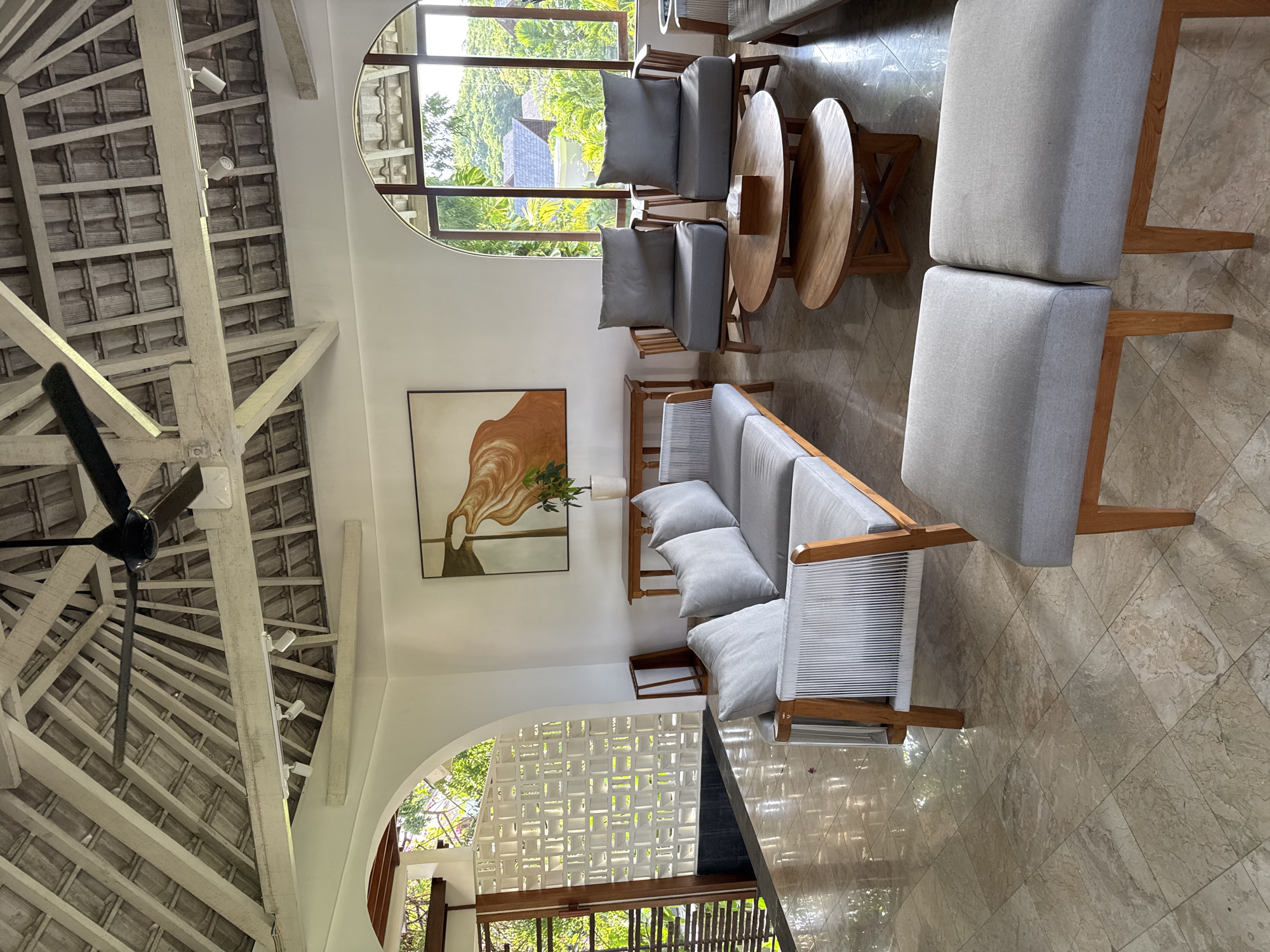The image shows an interior space in Kuta Selatan, Indonesia, featuring a seating arrangement with two armchairs and a sofa, all upholstered in light gray fabric with wooden frames. Two round wooden tables are placed between the armchairs. A large, abstract painting hangs on the wall. The room has a distinctive roof structure with exposed white beams and tiles. Natural light enters through a large arched window and a patterned window wall. The floor is tiled in a beige marble pattern. A ceiling fan is visible on the left side of the frame. No people are present in the image.