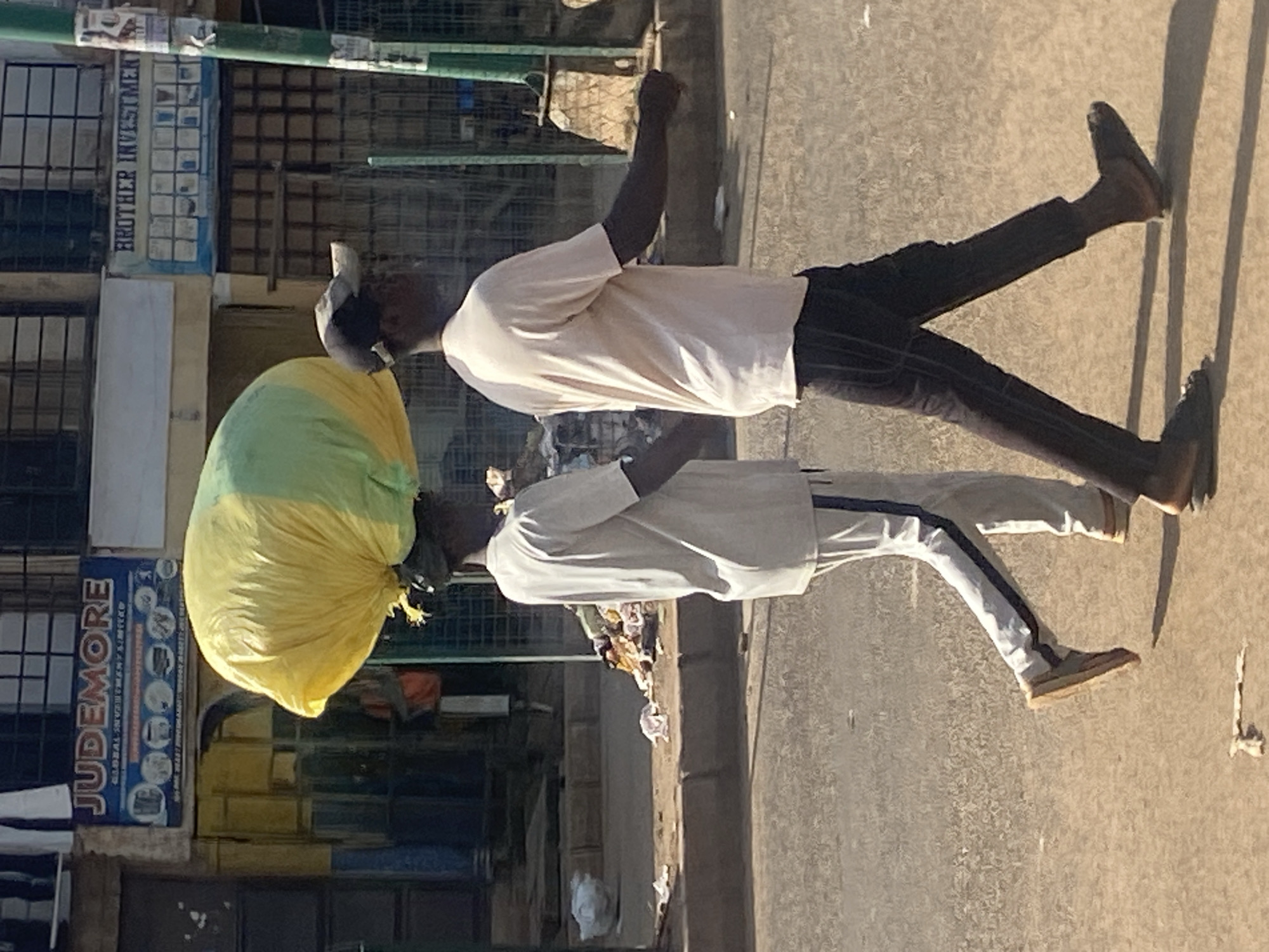 Two men are walking on a street in Bukuru, Nigeria. The man on the left is carrying a large yellow and green bundle on his head. He is wearing a light-colored t-shirt and pants with a blue stripe down the side. The man on the right is wearing a light-colored t-shirt and dark pants. The street is paved and there is some trash on the side of the road. In the background, there are buildings with signs. One sign reads "JUDEMORE" and another reads "BROTHER INVESTMENT". The scene appears to be a busy marketplace or commercial area.