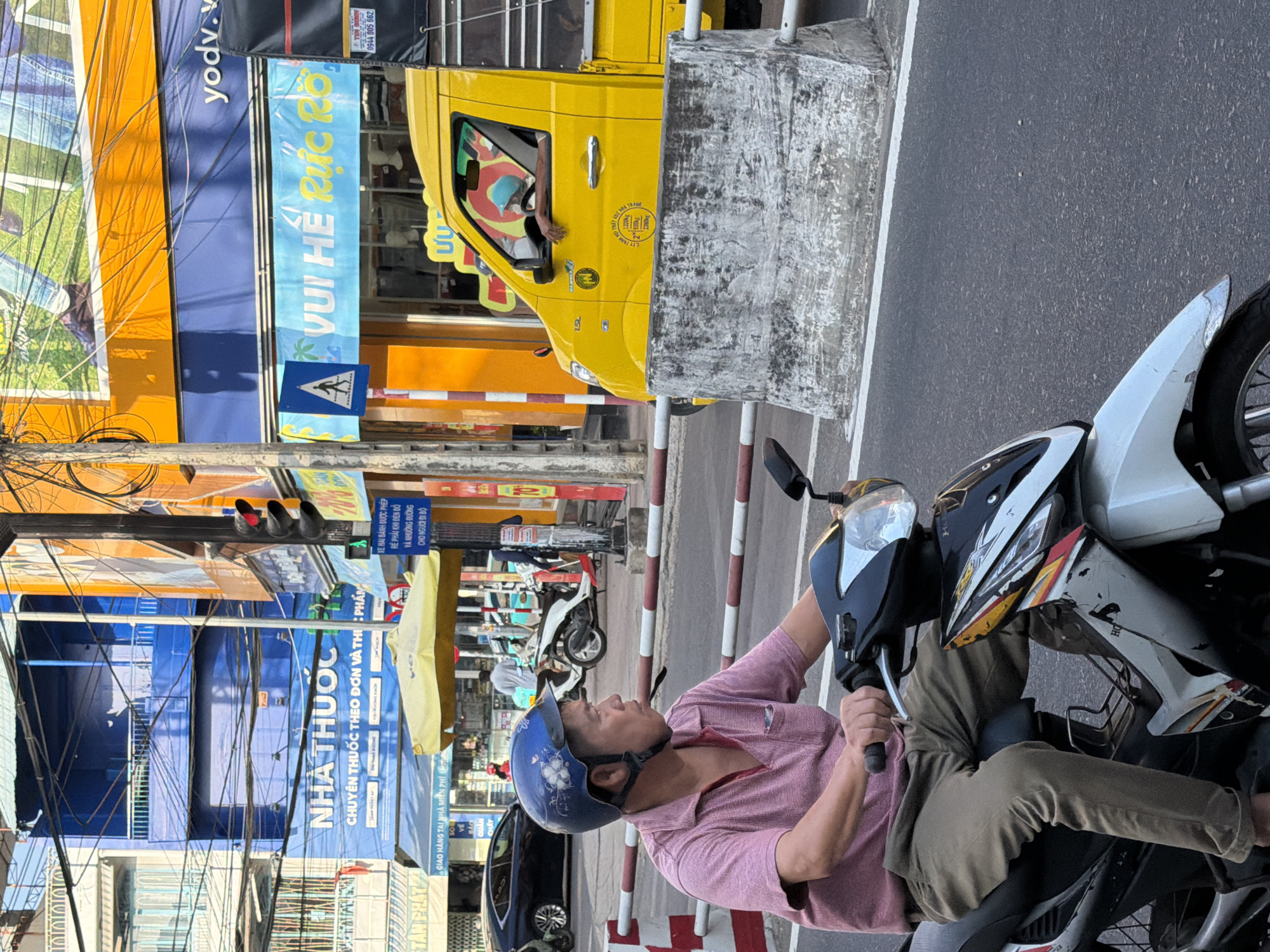 A man wearing a blue helmet and a pink polo shirt rides a white and black motorcycle on a city street in Nha Trang, Vietnam. He is looking to his right, with his hands on the handlebars. The street is paved and has a white line on the right side, separating it from a concrete barrier. In the background, there are several buildings, including a blue building with Vietnamese text that reads "NHÀ THUỐC" (pharmacy) and a yellow building with orange accents. A yellow truck is parked to the right of the motorcycle, and a traffic light is visible on the left side of the frame. The sky is bright and sunny.