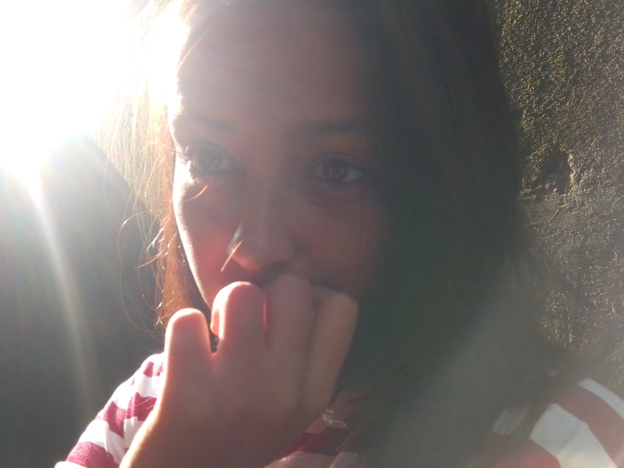 A close-up, low-angle shot shows a person's face partially obscured by bright sunlight and their hand. The person, with dark hair and brown eyes, is wearing a red and white striped shirt. Their right hand is raised, with fingers near their mouth. The background consists of a dark, textured wall on the right and a very bright, sunlit area on the left, creating a strong contrast. The image appears to be taken outdoors, possibly in a public area within Siliguri, India, given the bright sunlight and textured wall which could be a building exterior or natural rock formation.