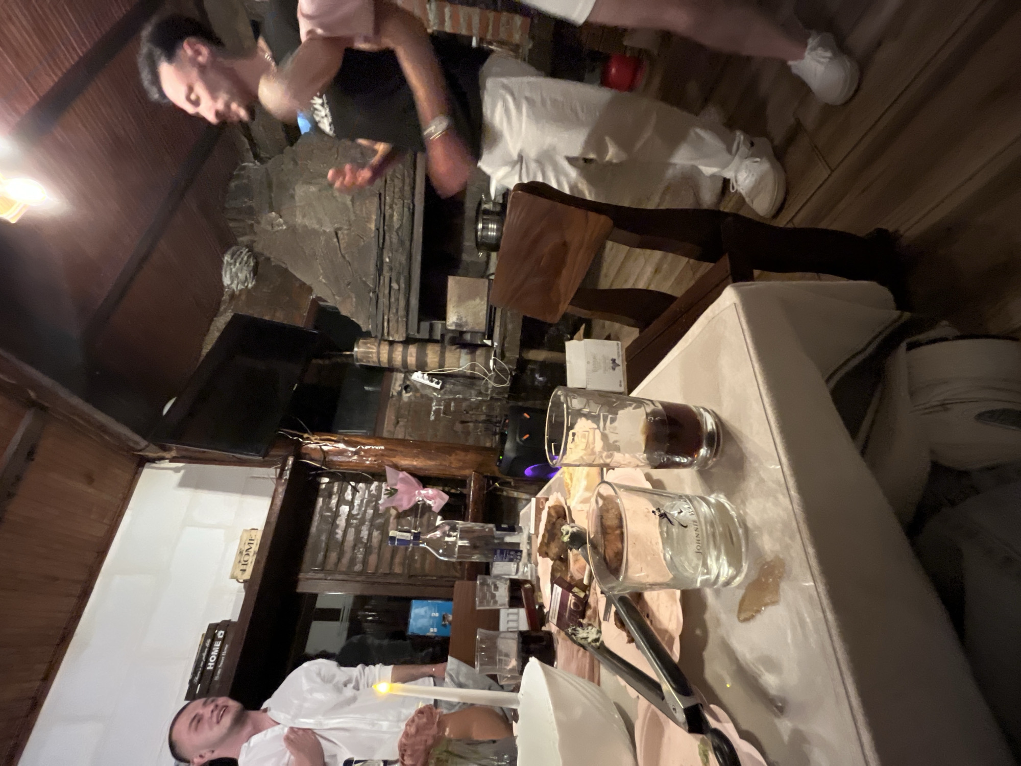 The image depicts an indoor dining scene, possibly in Presoka, Bulgaria. Two men are visible, one wearing a black t-shirt and white pants, the other's attire is partially obscured. A partially visible man is at the edge of the frame. A set table dominates the foreground, laden with glasses, bottles, and plates of food. The room features wooden floors and walls. Behind the table, a fireplace with stone accents can be seen, above which is mounted a flat-screen TV. Books and other objects are stacked on a shelf.