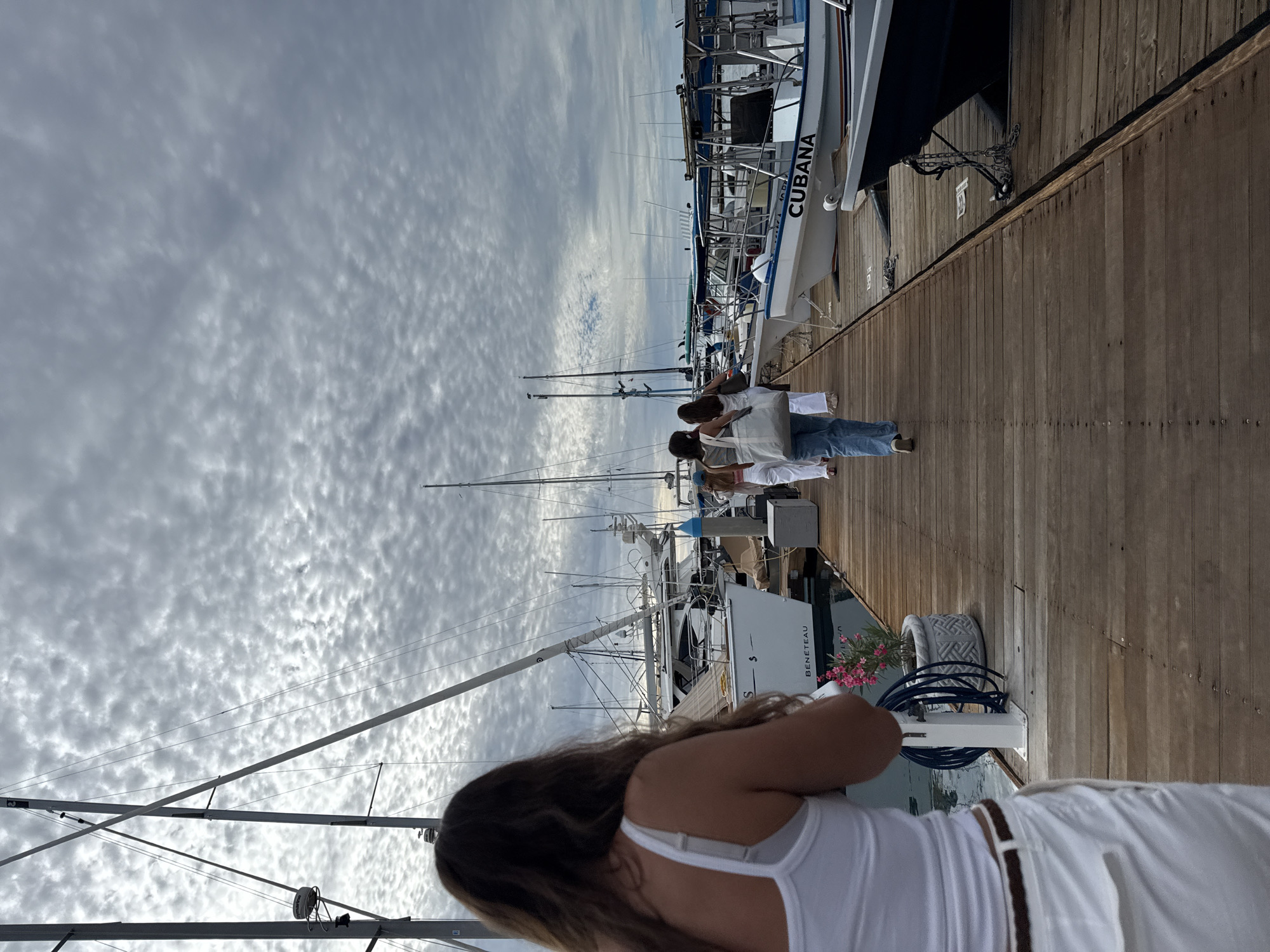 Here's a description of the scene:
The image shows a pier in La Paz, Mexico, filled with several sailboats. The perspective is oriented along the length of the pier. A group of people are walking along the pier. In the foreground, a woman with long hair is visible, wearing a white tank top and white pants with a brown belt. The sky is overcast with a layer of cloud cover. The sailboat "Cubana" is seen docked on the pier. A "Beneteau" sailboat is also visible. A gray flowerpot with pink flowers sits on the side of the pier. The pier is constructed of wooden planks.