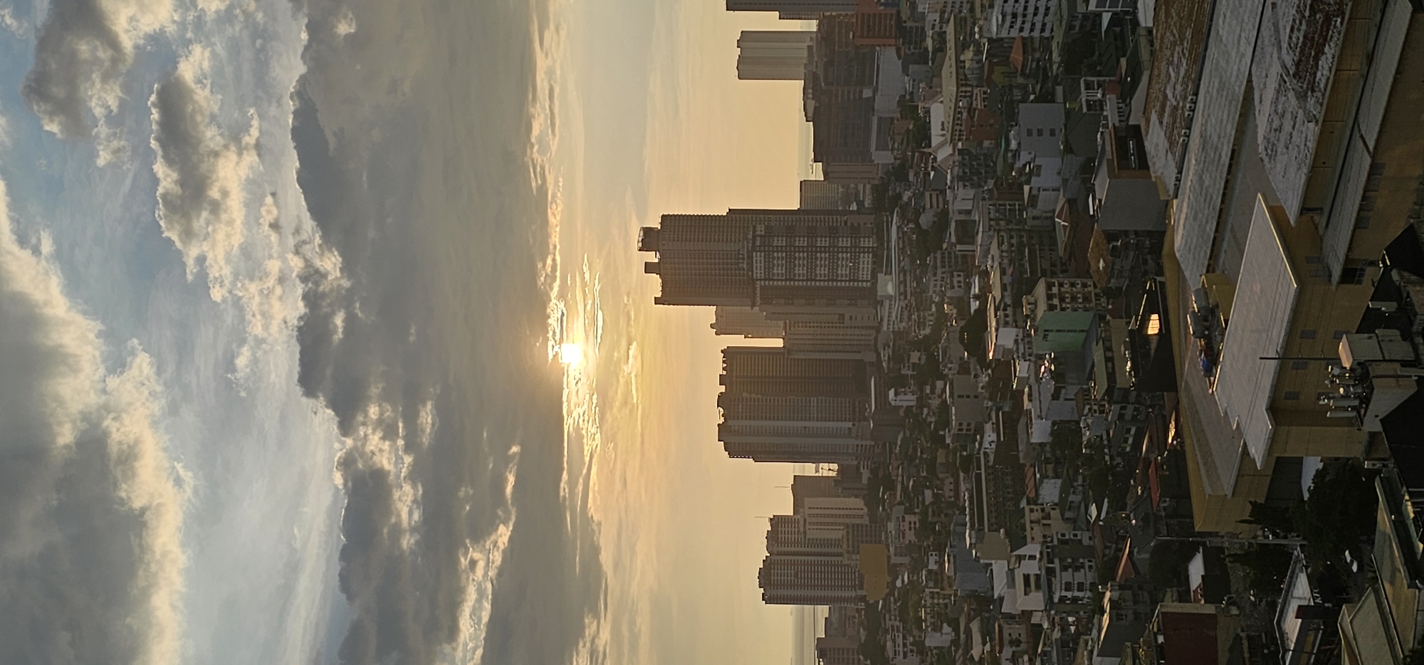 This is a vertical, medium-distance, eye-level shot of the Makati skyline during what appears to be a sunrise or sunset, judging by the warm lighting and scattered clouds. The sun is visible as a bright orb peeking through a gap in the clouds, casting a golden hue across the sky and the upper portions of the buildings. The scene is dominated by a dense collection of high-rise buildings and lower-rise structures, indicative of an urban environment. Some of the taller buildings feature intricate window patterns and varied architectural designs, suggesting a mix of residential and commercial spaces. The lower sections of the image show a more varied urban landscape with rooftops, some appearing to be flat and others with various structures like water tanks or ventilation units. The overall impression is a bustling cityscape under a dramatic sky.