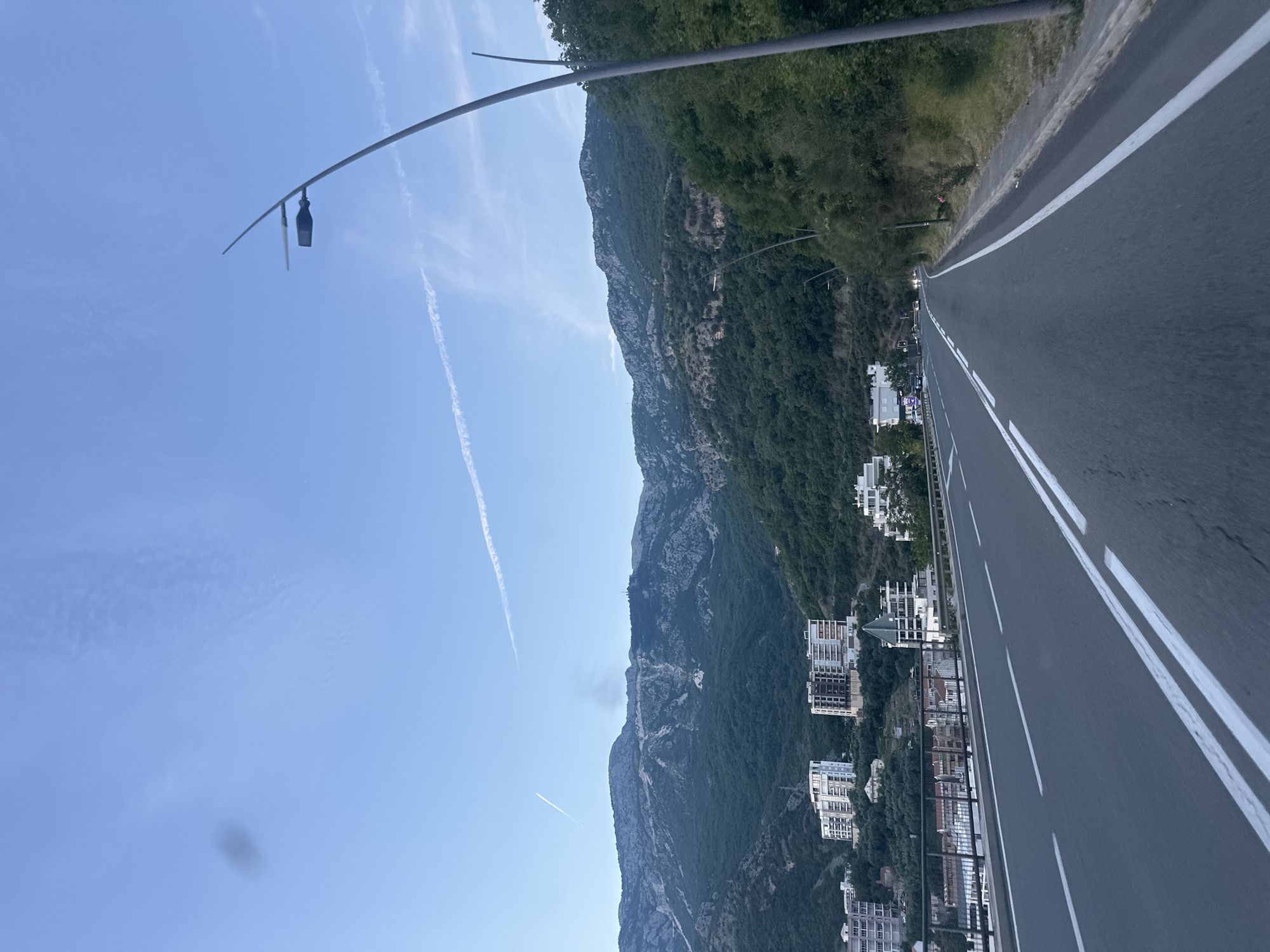 The image displays a vibrant daytime scene, capturing a scenic coastal road winding alongside a lush mountainous landscape under a clear blue sky.
In the foreground, a paved asphalt road with prominent white double solid lines stretches from the bottom right towards the upper left. A metal guardrail or fence runs along the left edge of the road, separating it from the adjacent terrain.
Beyond the road and to the left, a majestic mountain range dominates the midground. Its steep slopes are densely covered with rich green vegetation, punctuated by areas of exposed grey rock. Nestled at the base of the mountain and between it and the road are numerous modern, multi-story buildings, primarily white or light-colored, suggesting a developed town or resort area.
The background features a bright, clear blue sky, marked by several faint white contrails. A lone street light with a curved arm extends into the upper-middle of the frame, reaching over the road.
The scene is bathed in bright daylight, indicating a sunny day with excellent visibility. The combination of steep, verdant mountains, coastal development, and a well-maintained road is characteristic of the Rafailovići region in Montenegro. There is no visible text in the image.