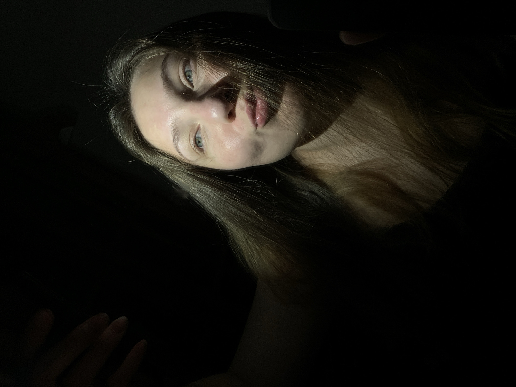 A young woman is captured in a selfie taken in a dark environment, possibly an indoor space in the city of Chaikovsky, Russia. The lighting is dramatic, with a strong light source illuminating the left side of her face and hair, creating stark shadows. She has light brown hair that appears to be slightly wind-blown across her face, obscuring parts of her features. Her eyes are open and looking to her left, and her lips are pursed. A hand is visible in the upper left corner, appearing to be holding a phone or camera for the selfie. The background is almost entirely black, offering no discernible details of the location or surroundings.