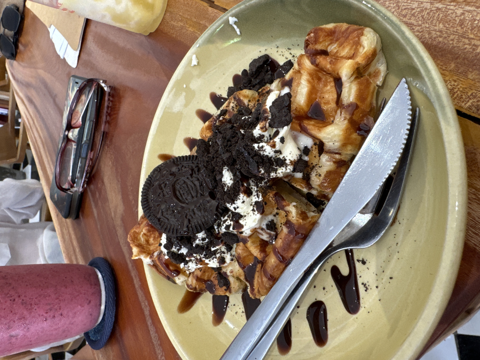 An overhead, close-up shot captures a decadent dessert and other personal items on a wooden table. The central focus is a light yellow-green ceramic plate holding what appears to be croffles or waffles. They are generously topped with white whipped cream, drizzled with dark chocolate syrup, and sprinkled with crushed dark chocolate cookies. A whole dark chocolate sandwich cookie, with the word "OREO" embossed on its surface, rests on the cream. A silver serrated knife and a matching fork are placed on the plate, ready for use.
To the left of the plate, a tall glass of a pink or reddish smoothie, sitting on a round blue coaster, is partially visible. Further up on the wooden surface, a pair of black sunglasses and a pair of red-framed reading glasses are casually placed. A smartphone is partly obscured beneath the reading glasses. A crumpled piece of paper, possibly a napkin or menu, is also visible. The wooden table serves as the backdrop for this scene, suggesting a casual dining setting like a cafe or restaurant in Phu Thuy Ward, Vietnam. No people are visible, and the bright lighting suggests it is daytime.