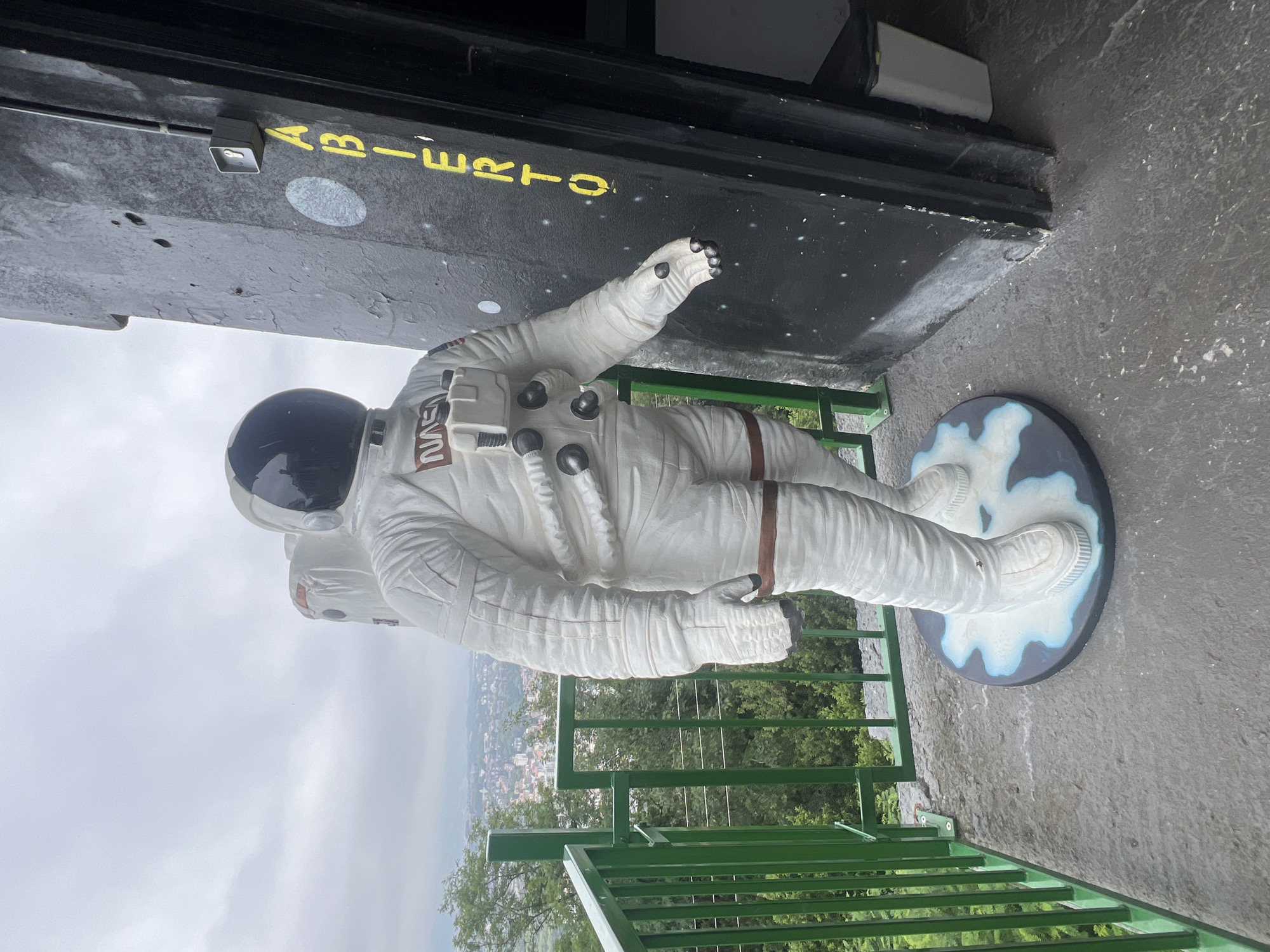 A statue of an astronaut stands on a concrete balcony next to a black wall with yellow lettering that reads "ABIERTO." The astronaut is wearing a white spacesuit with a black helmet and a NASA logo on the chest. Behind the astronaut, a green railing overlooks a city with buildings nestled among lush green trees and hills. The sky is overcast. A white trash can is visible to the right of the black wall. The scene appears to be outdoors in a city with a view of a landscape.