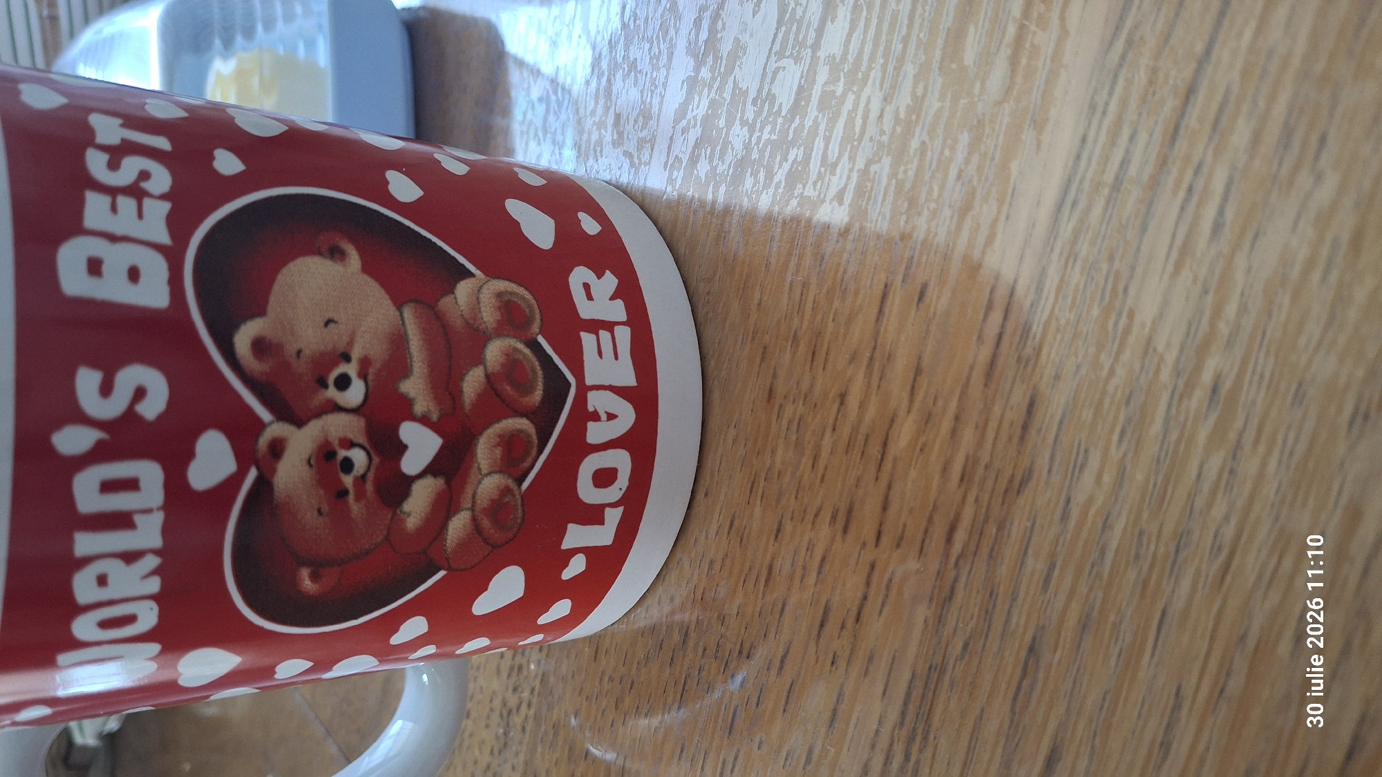 The image shows a red mug with white polka dots and a heart graphic depicting two teddy bears. The text "WORLD'S BEST LOVER" is visible on the mug. The mug is placed on a wooden table. A blue and white object, possibly a container, is partially visible in the background. The bottom right corner of the image has a timestamp: "30 iulie 2026 11:10". There are no people visible in the image. The scene appears to be indoors, given the lighting and the presence of a table and mug. The location is identified as Ialoveni, Moldova, though this information is not visually present in the image itself.