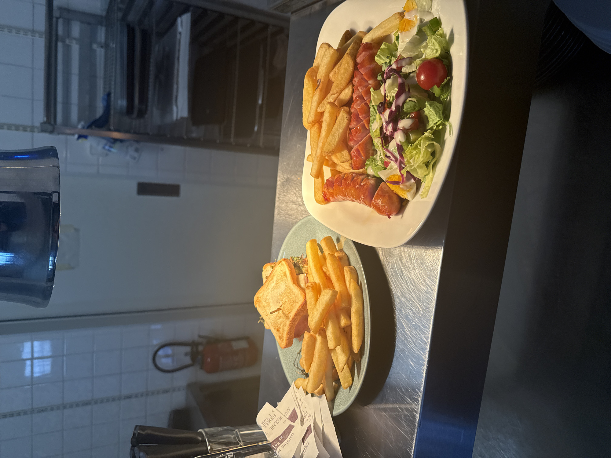 Two plates of food are presented on a metallic surface, likely a counter in a restaurant or catering establishment. The plate in the foreground contains a sandwich, cut diagonally and served with a generous portion of french fries. The sandwich appears to be toasted, with a filling visible. Below the plate, several crumpled receipts or order slips are partially visible. The plate in the background holds a salad with lettuce, red cabbage, cherry tomatoes, hard-boiled egg slices, and two sausages, accompanied by french fries. The lighting suggests it is daytime, possibly indoors, with some natural light filtering in, as evidenced by the reflections on the metallic surfaces. The overall scene is functional and practical, focused on the food presentation. The location context provided is Rivesaltes, France, but this is not directly inferable from the visual information alone. There is visible text on the receipts, but it is too blurry to be legible.