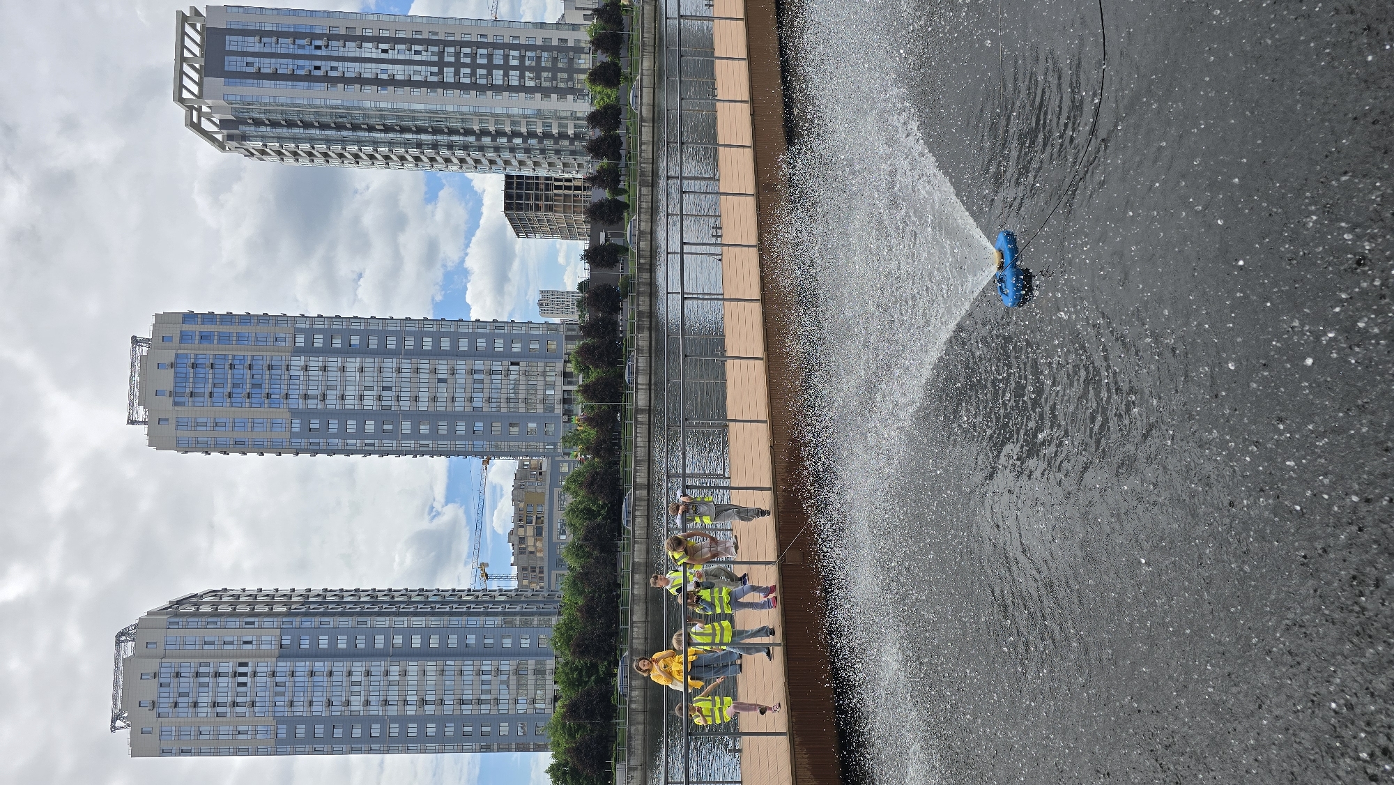 This is a photograph taken on a cloudy day at a waterfront in Kyiv, Ukraine. In the background, three modern, tall residential buildings with blue-tinted windows are visible, two of which appear to be under construction with cranes. A body of water, likely a river or canal, separates the foreground from the background.
In the mid-ground, a group of children, wearing bright yellow safety vests, stand on a wooden pier. One adult, also in a yellow top, is with them. The children appear to be looking out at the water.
The foreground of the image is dominated by a water fountain spraying water in a wide arc. A small, blue, toy-like object, possibly a remote-controlled boat, is visible in the water near the fountain, connected by thin wires. The water surface is agitated by the fountain's spray, with numerous droplets visible. The overall scene suggests a recreational area in a city park or waterfront development.