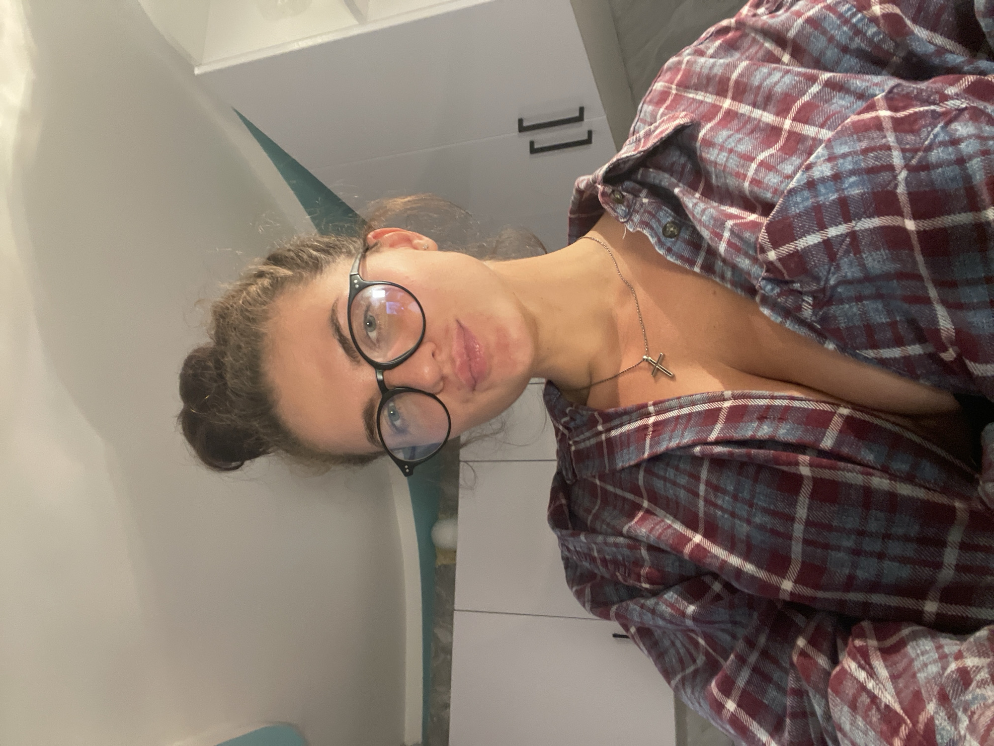 A young woman with her hair in a messy bun and wearing glasses, a plaid shirt, and a necklace with a cross, looks into the camera. She is in a kitchen.