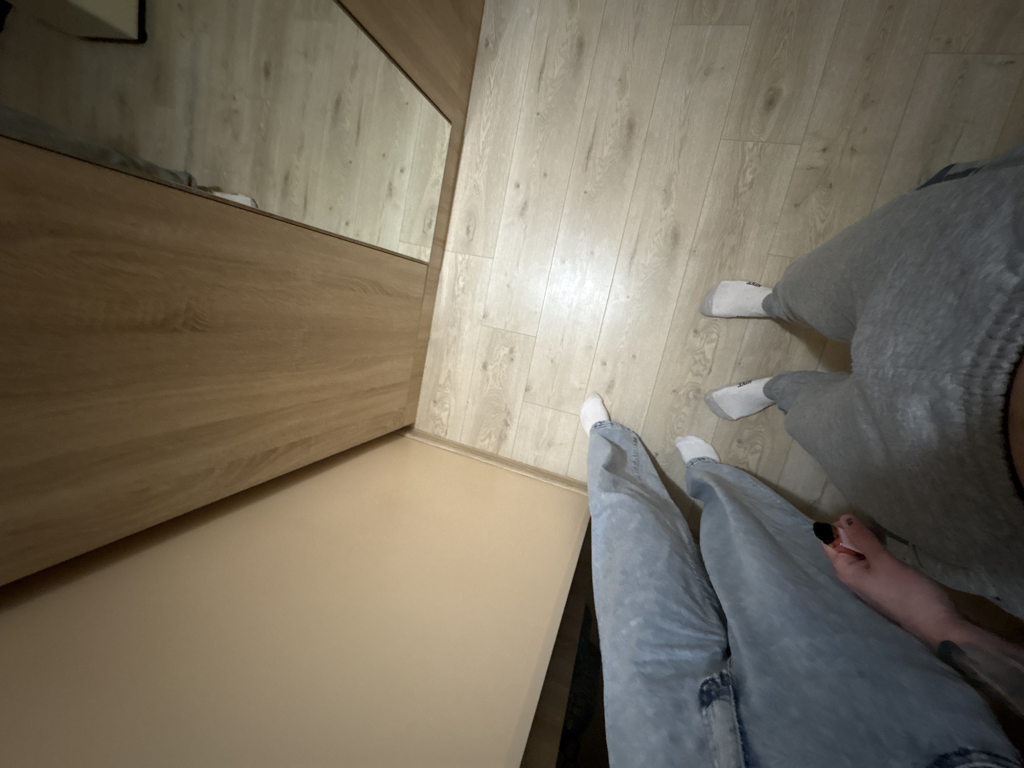 A high-angle shot shows two pairs of legs in what appears to be an interior setting. One pair is wearing light-wash denim jeans with white socks, and the other is wearing gray sweatpants with white socks. The floor is light-colored wood laminate. A portion of a light brown wooden cabinet or wall is visible on the left, along with a mirror reflecting the floor. A light beige-painted wall is visible along the bottom and left side of the frame. The location is Chaikovsky, Russia.