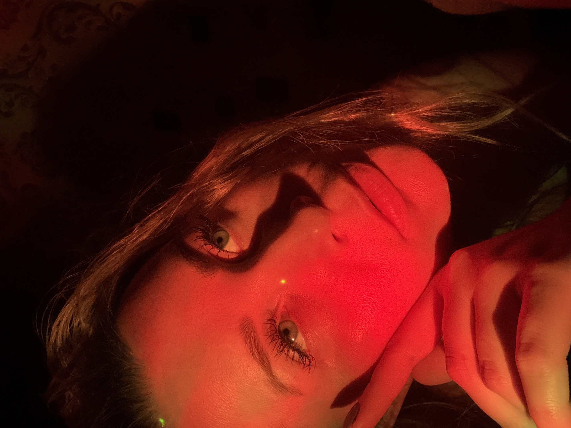 The image portrays a young Caucasian woman situated in the city of Veliky Novgorod, Russia, illuminated predominantly by a red light source emanating from the viewer's left. Her face is centered, with her left hand positioned against her cheek and jawline. A small green laser dot is visible on her upper forehead, and a distinct yellow-orange laser dot is present on the bridge of her nose. Her eyes, featuring green irises and pronounced eyelashes, are directed towards the camera with a slight upward gaze. The right side of her face and the background are enveloped in deep shadow, although subtle patterns, indicative of wallpaper or a textured surface, can be discerned in the upper right background, suggesting an indoor setting. Her long, light brown hair is visible along the right side of her head and extends over her shoulder.