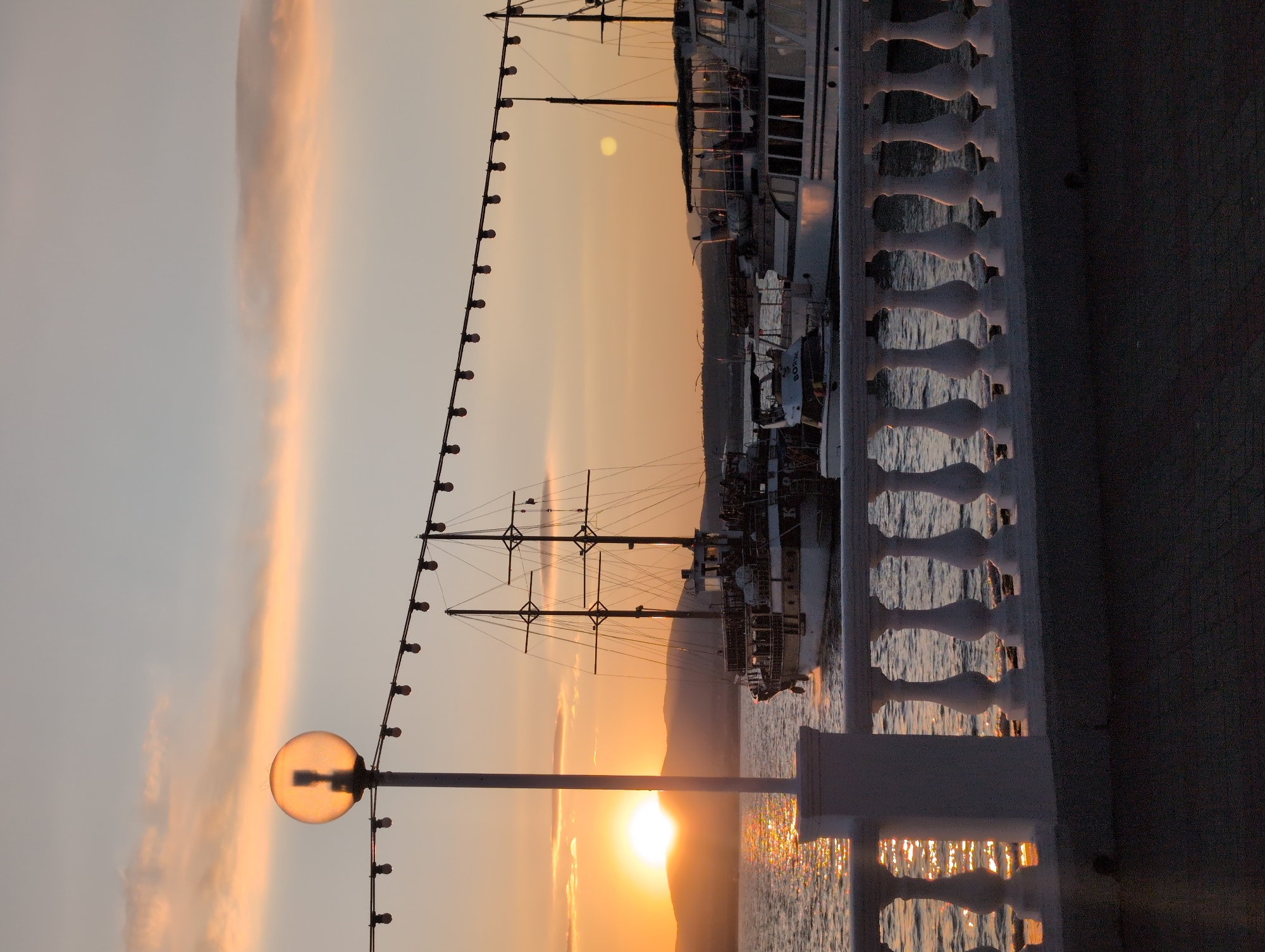 The image depicts a sunset scene at the Gelendzhik embankment in Russia. A body of water, likely the Black Sea, is visible, with its surface reflecting the warm hues of the setting sun. Several boats, including a large sailing vessel with multiple masts and several motorboats, are docked or anchored in the water. In the foreground, a white balustrade runs horizontally across the frame, separating a paved walkway from the water. A lamppost with a spherical lamp and a string of lights extending outwards are also present, adding to the ambiance of the promenade. The sky is painted with soft orange, pink, and yellow tones, indicating the time of day. The overall atmosphere is tranquil and picturesque.